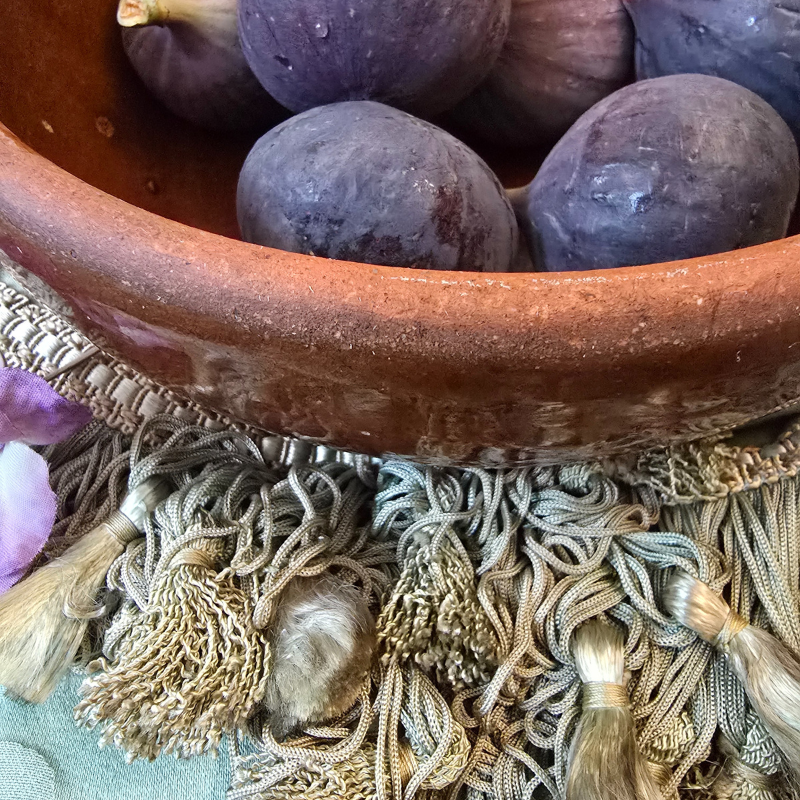 Rustic French tian bowl filled with deep purple figs, styled on an ornate tasselled textile for early autumn mood.