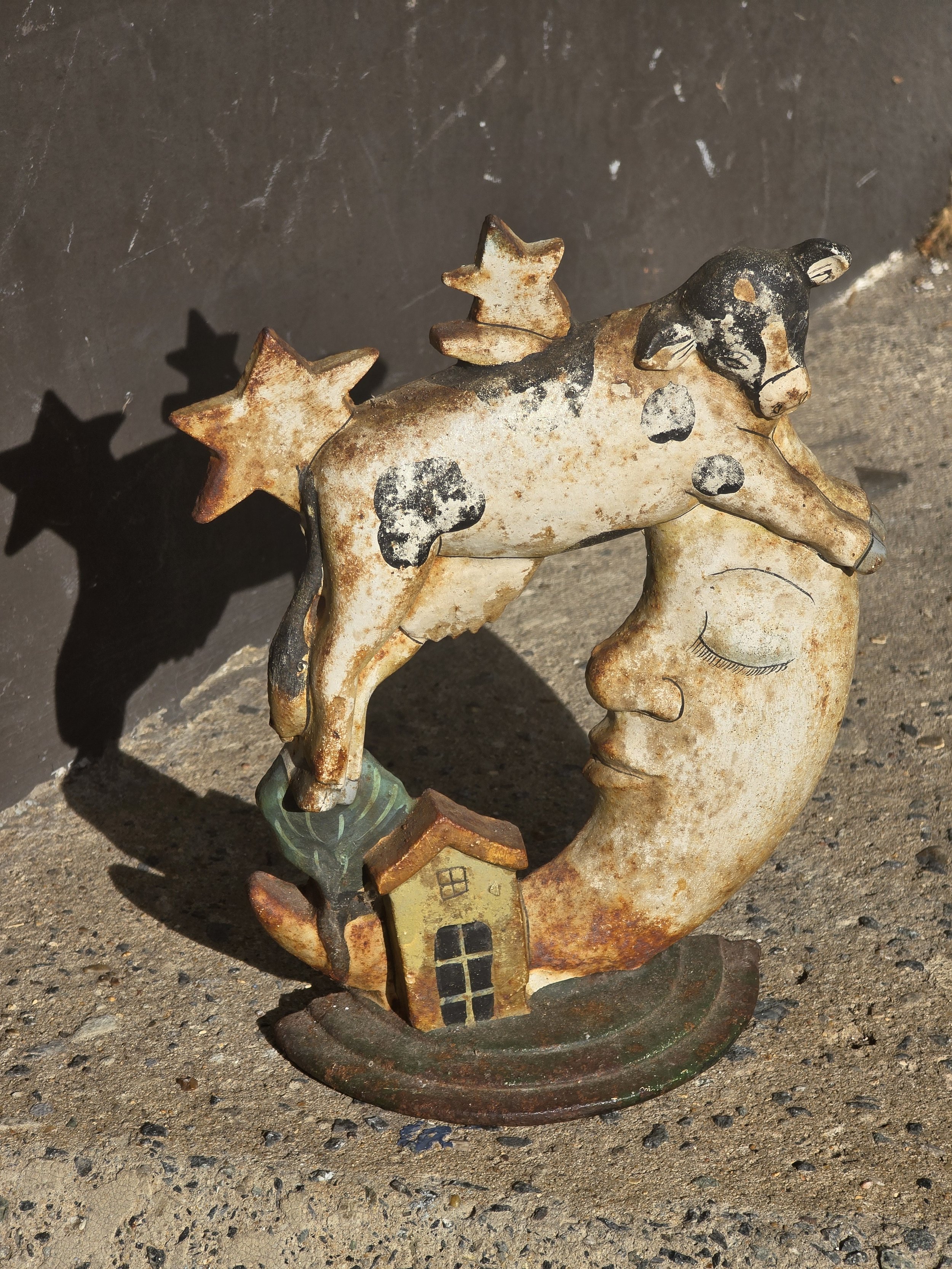 Timeworn vintage metal ornament depicting a black and white cow jumping over a crescent moon with stars and a tiny house.