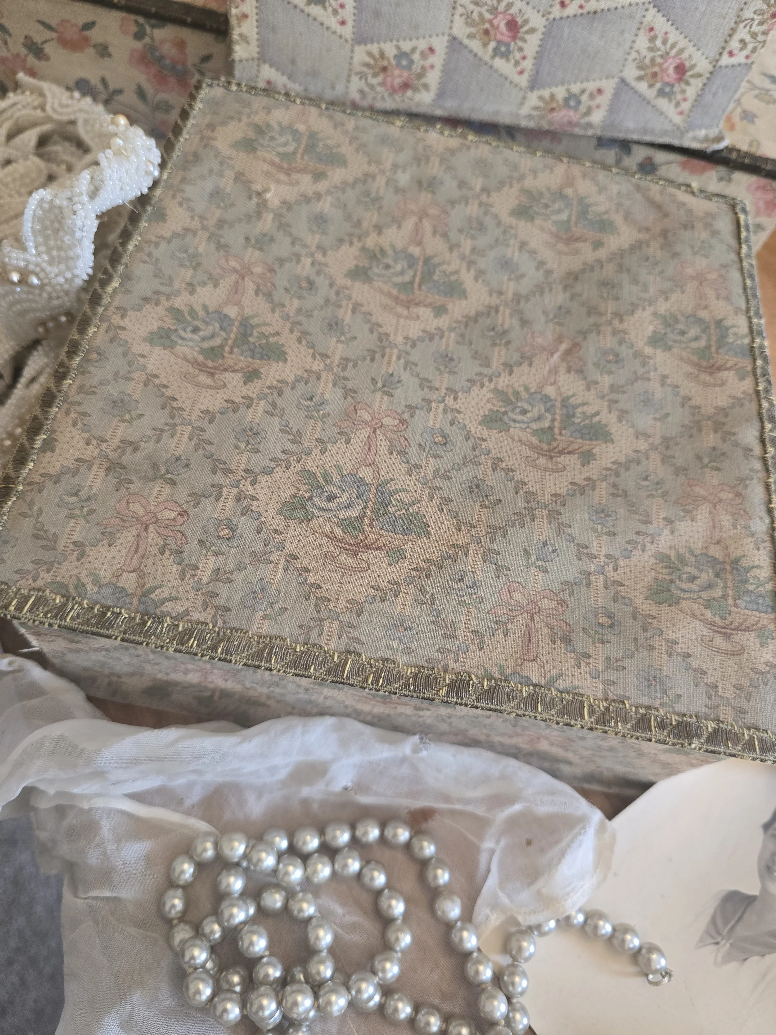 Antique French fabric box styled with vintage pearl beads and old textiles, showing the faded floral lid.