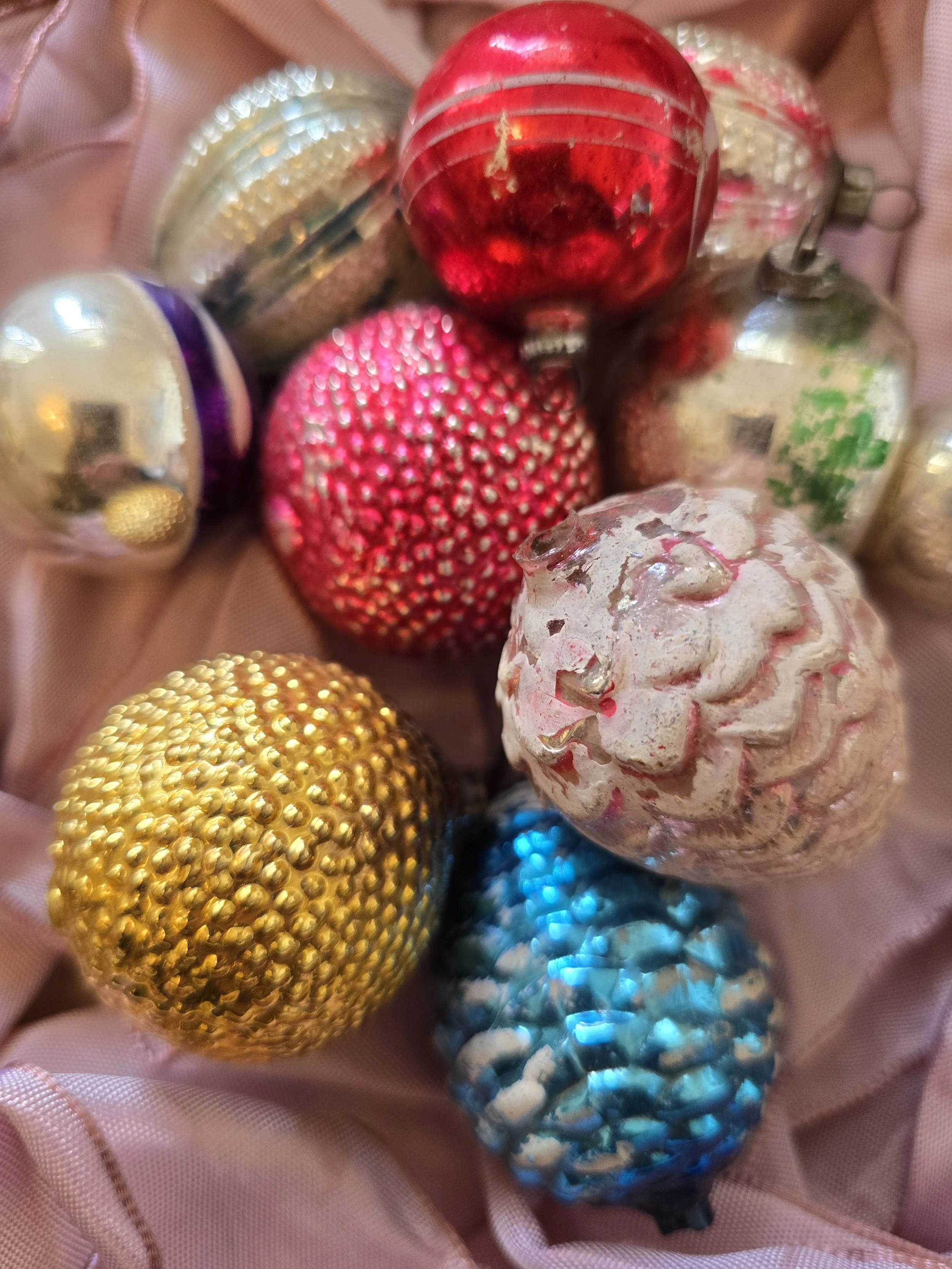 Vintage glass Christmas baubles with purple, red and silver painted designs arranged on pink satin fabric.