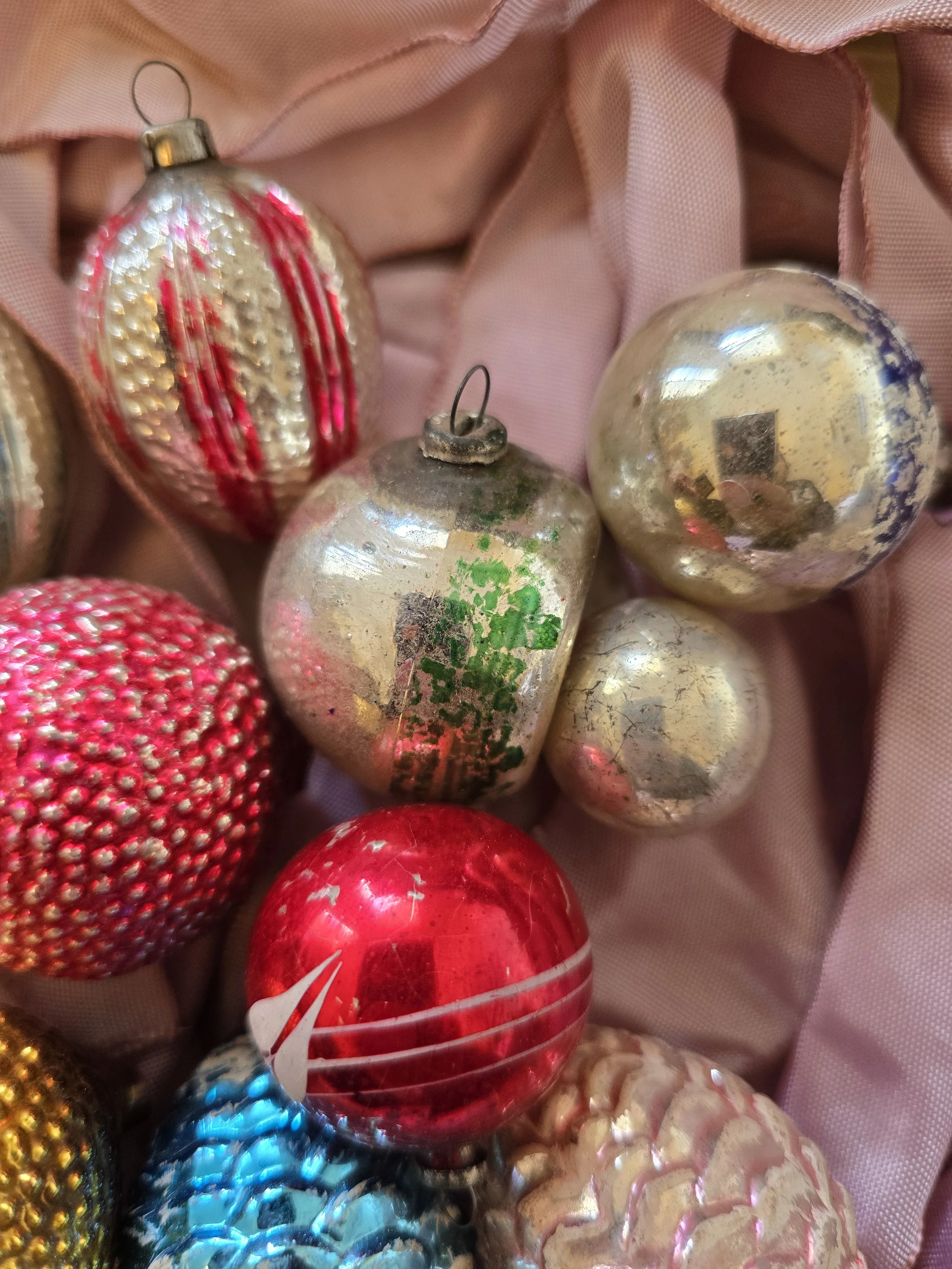 Collection of eleven colourful vintage glass Christmas baubles in pink, red, gold, silver, blue and purple, displayed on pink fabric.