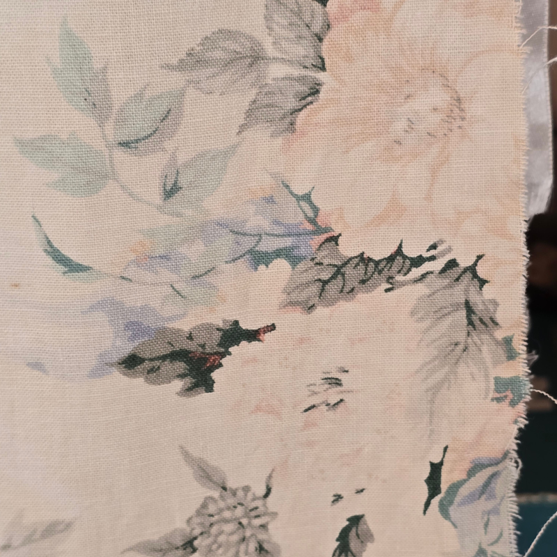 Close-up of a faded vintage floral fabric in soft blush, cream, pale blue and sage green tones. The worn textile shows frayed edges and age-softened colours, highlighting the beauty of timeworn cloth and gentle patina.