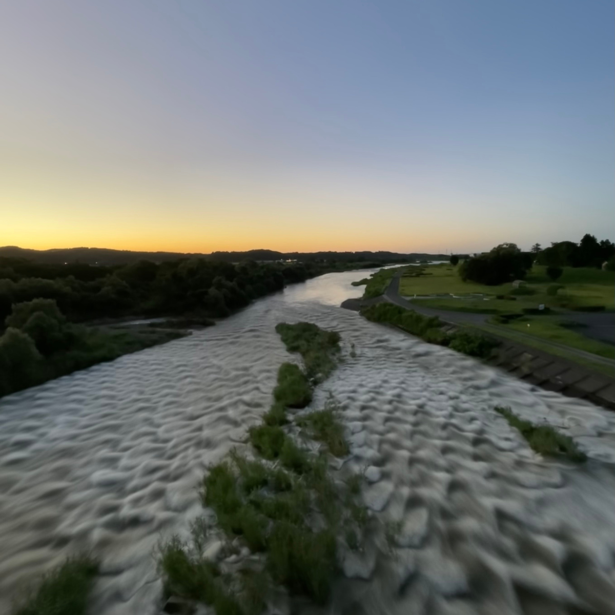 Seen from above at twilight, a broad river curves through the landscape as the sky softens from gold to blue. Islands of greenery divide the flowing water, creating a scene of quiet movement and reflection at the end of the day.