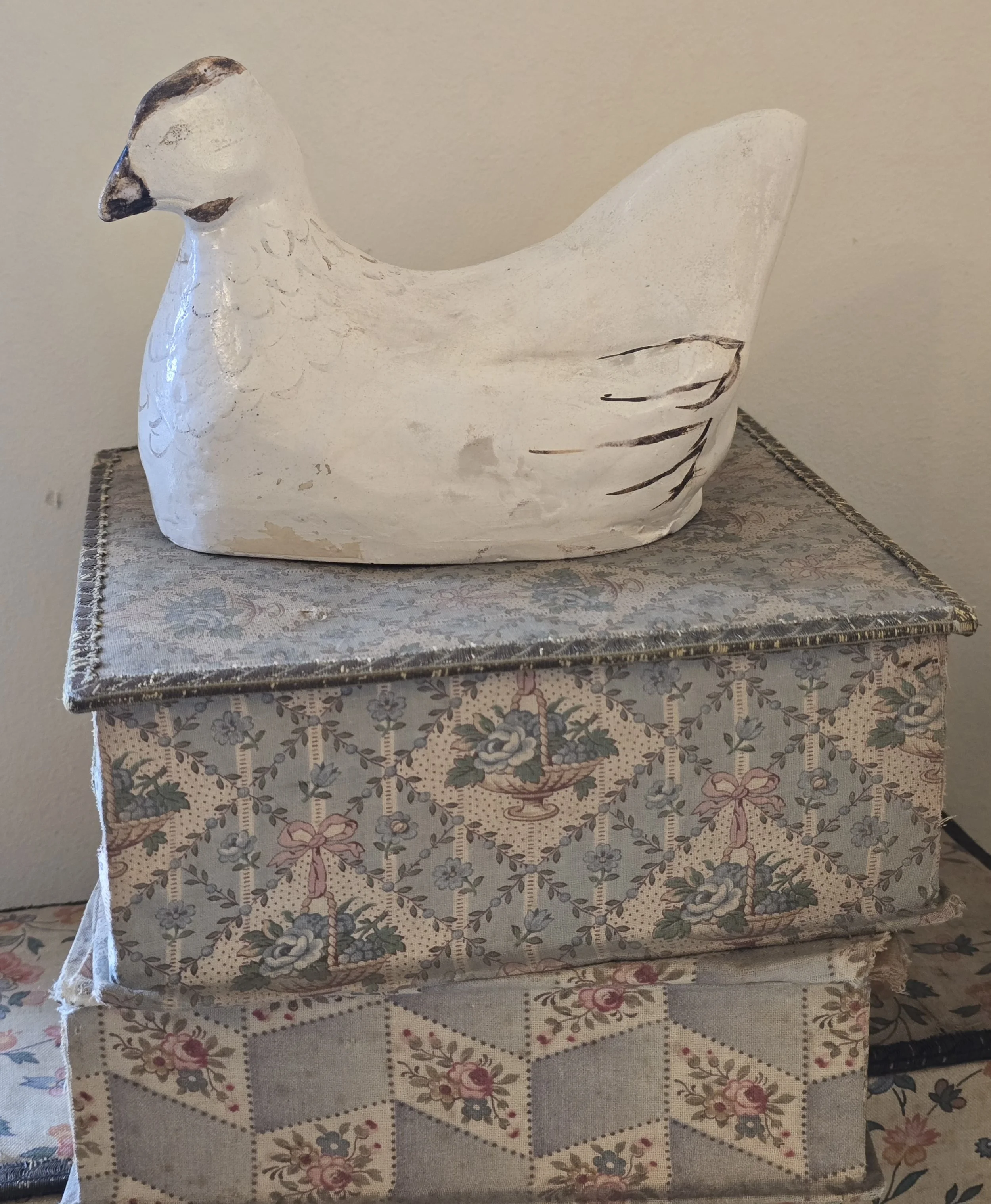 Old white painted hen ornament with worn black feather details, displayed on stacked antique fabric-covered boxes.