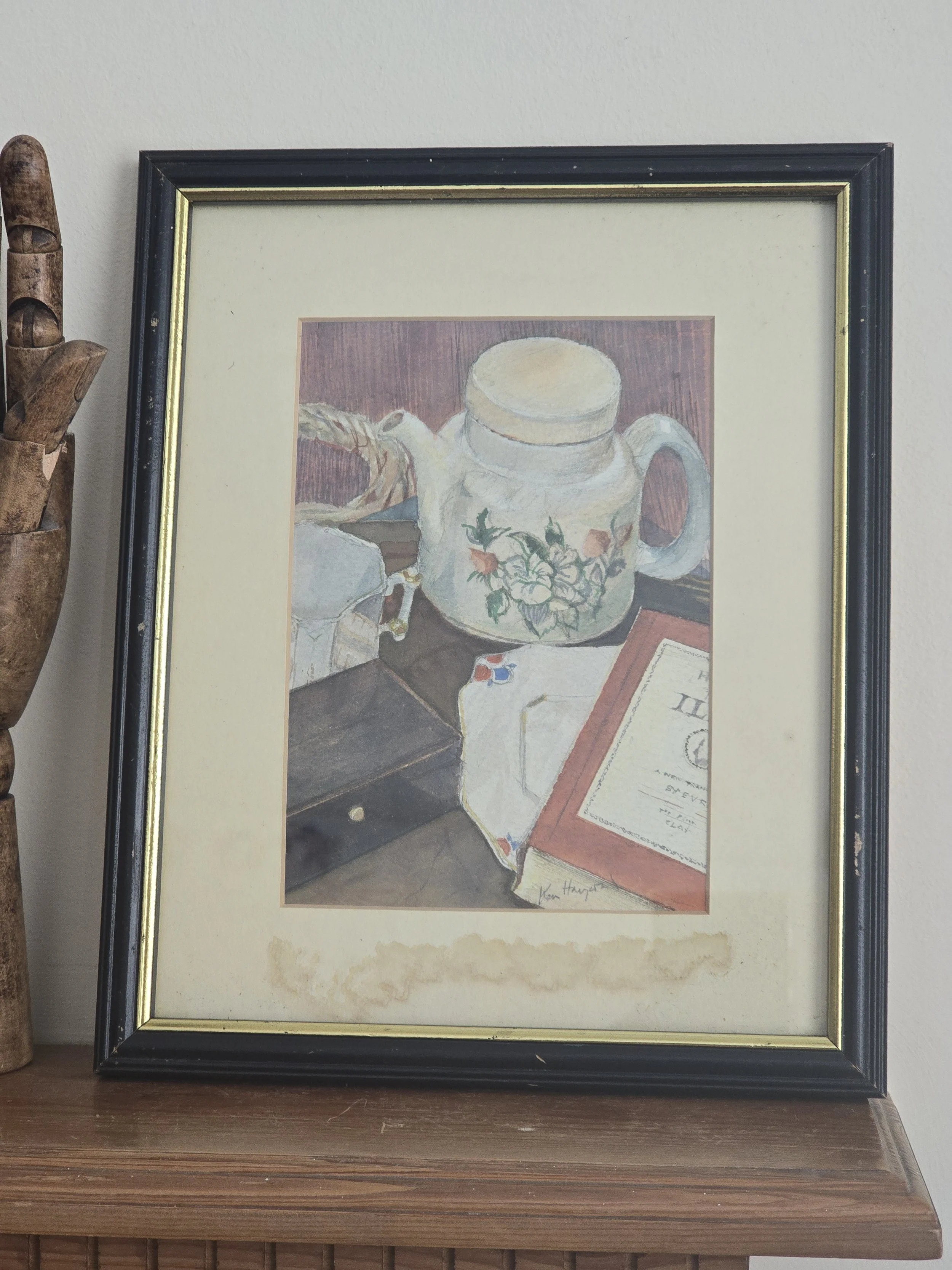 Signed original still life watercolour depicting a floral jug, folded textile and book in soft muted colours, presented in a vintage frame with aged mount.