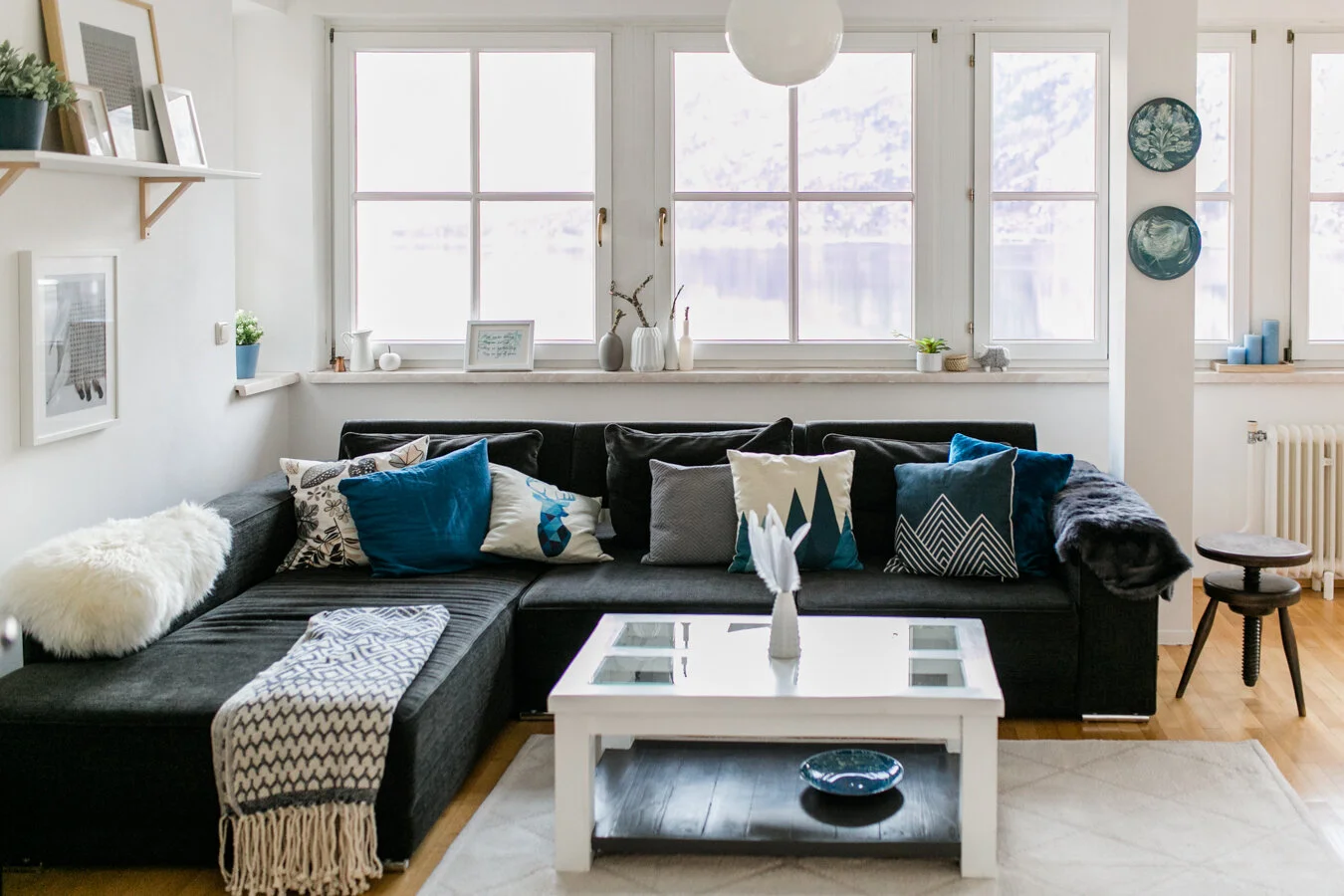 Gemütliches Wohnzimmer mit schwarzen Sofa, bunten Kissen, weißen Tisch und großen Fenstern, die viel Licht hereinlassen.