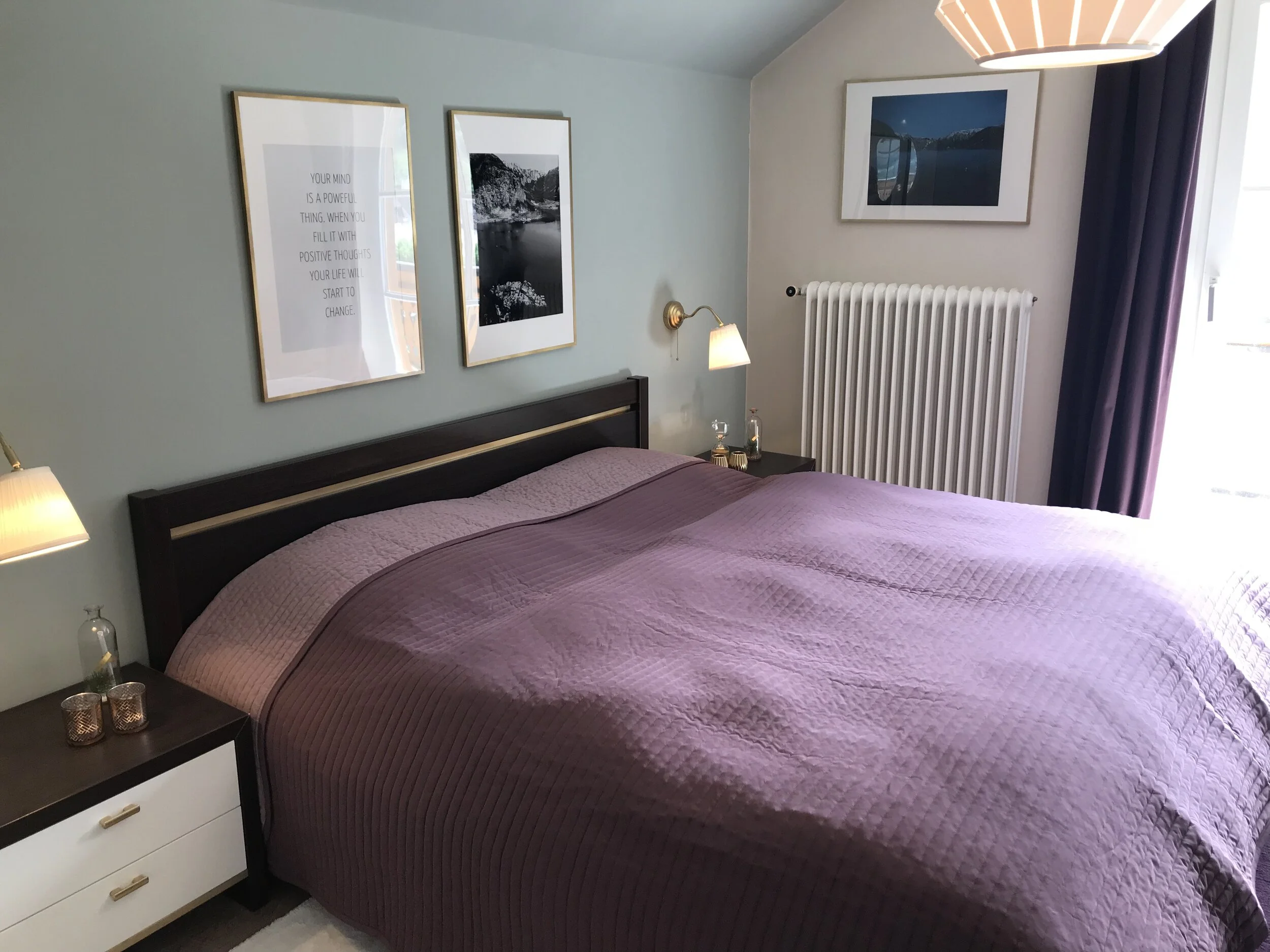 Bett mit lila Decke, Wandbilder, Nachttisch mit Kerzen, Wandlampe, Vorhänge, Fenster und Heizkörper in einem Schlafzimmer.