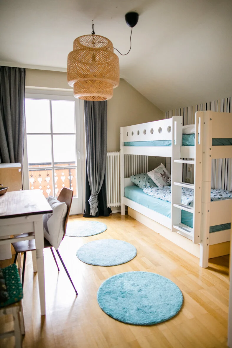 Ein Kinderzimmer mit einem weißen Etagenbett, einem Esstisch mit Stühlen und einem großen Fenster mit dunklen Vorhängen.
