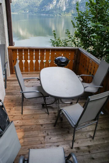 Sitzbereich auf einer Holzveranda mit Tisch, vier Stühlen, einem kleinen Grill und Blick auf einen See und Bäume.