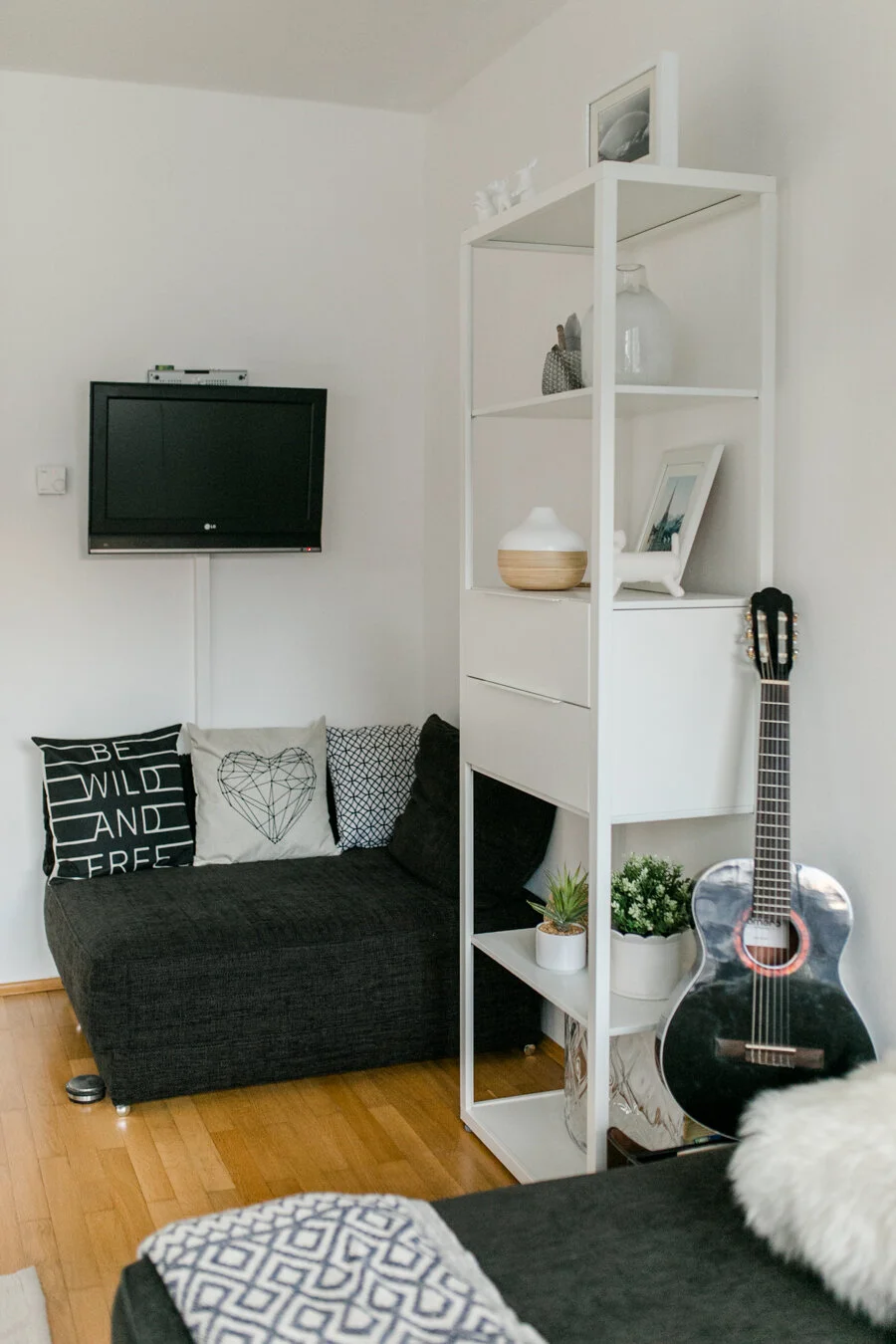 Wohnzimmer mit schwarzer Couch, weißen Kissen, einem flachen schwarzen Fernseher, einem weißen Regal mit Dekorationsgegenständen, Pflanzen und einer Gitarre