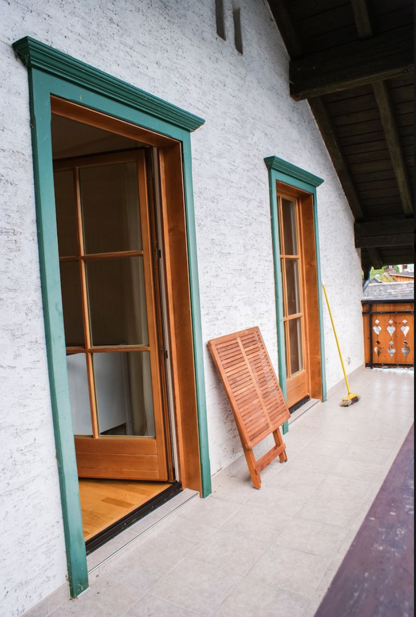 Balkon mit zwei Fenster-Türen mit grünen Rahmen, ein Holzvorhang, ein Besen und eine dekorierte Holzbrüstung.