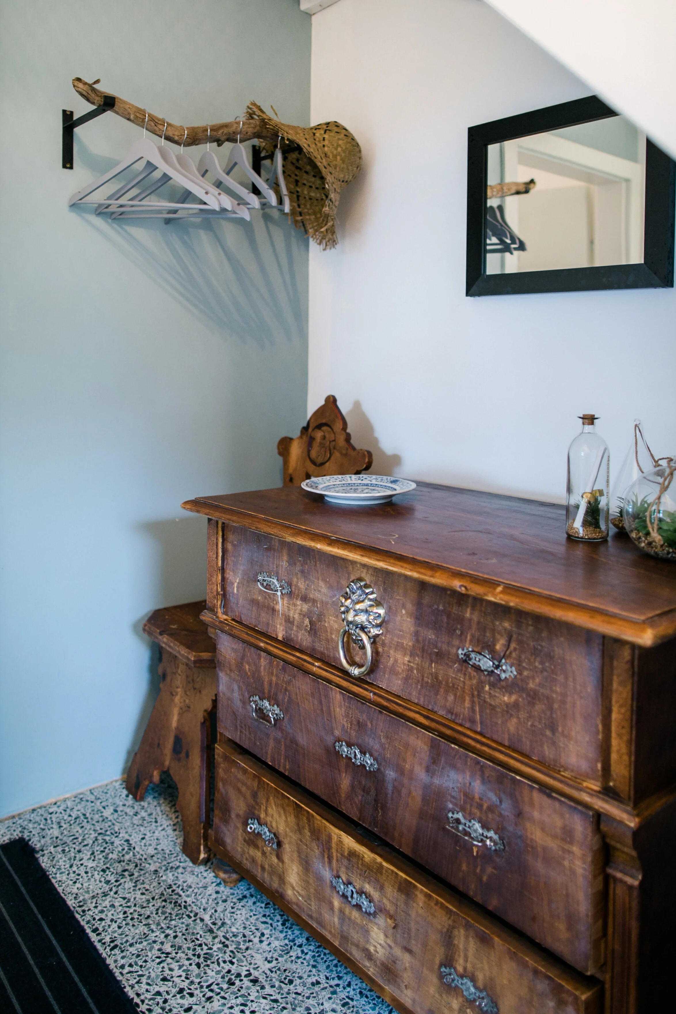 Alter Holzmöbel und Dekorationen in einer Wohnung, darunter ein antiker Holzschrank, ein Wandspiegel, ein Wandhaken mit Kleiderbügeln, eine Kette, eine Sessellehne, eine geplante Dekoplatte, mehrere Glasvasen mit Pflanzen und ein Fieck.