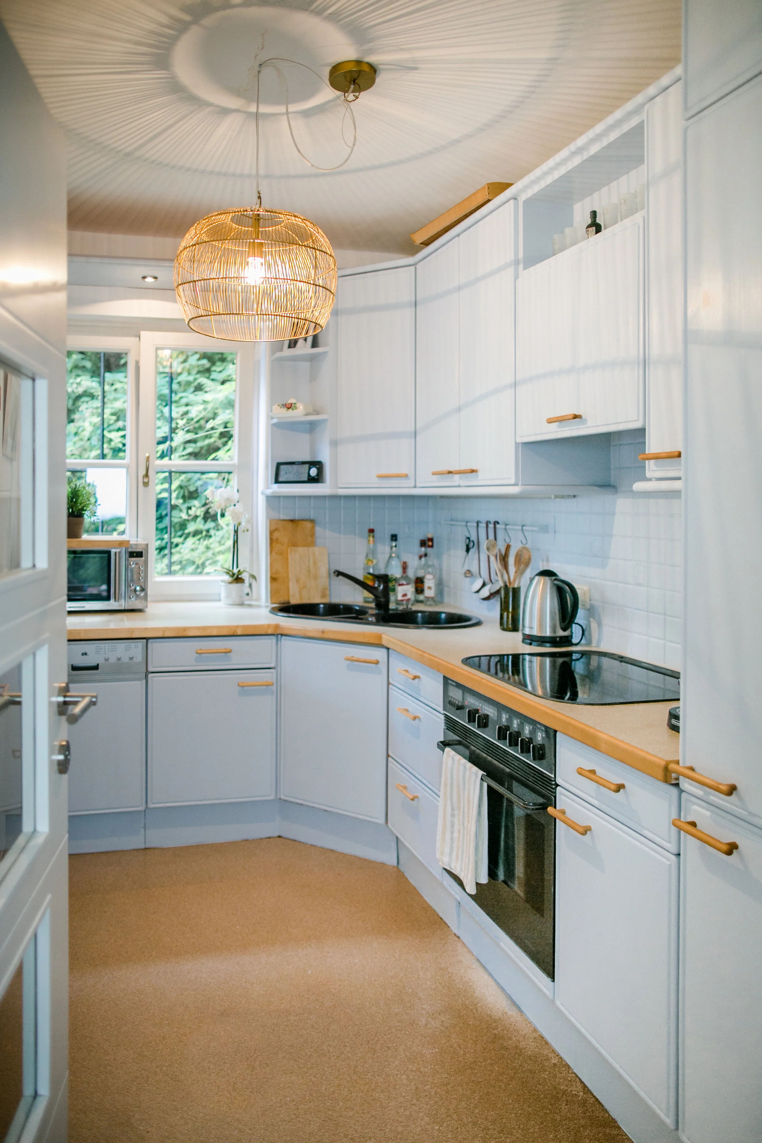 Küche mit weißen Schränken, Holzkanten, Herd, Backofen, Wasserkocher, Mikrowelle, Fenster mit Ausblick auf grüne Bäume, dekoratives Hängeleuchte, beige Teppichboden.