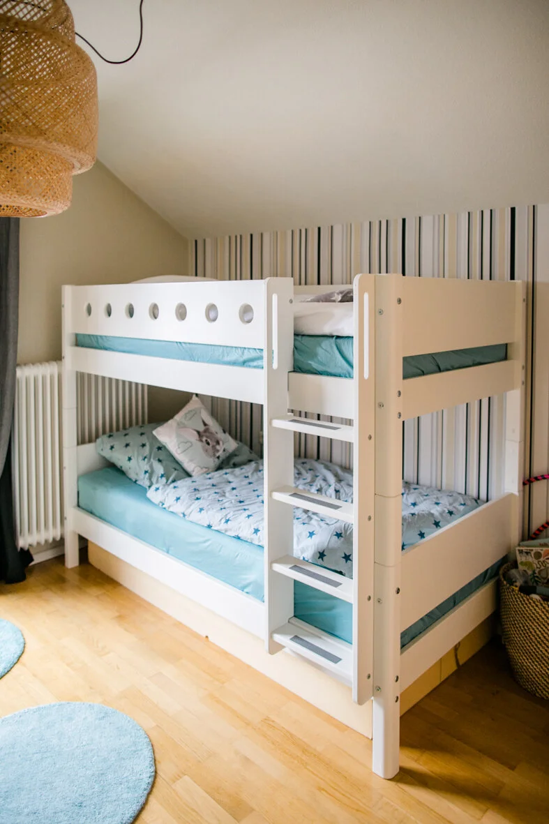 Hochbett mit zwei Betten, das im Kinderzimmer und Gästezimmer steht. Das Bett hat eine Leiter und ist vor einer Wand mit vertikalen Streifen. Es gibt Kissen und Bettwäsche in Blau und mit Tiermotiven.