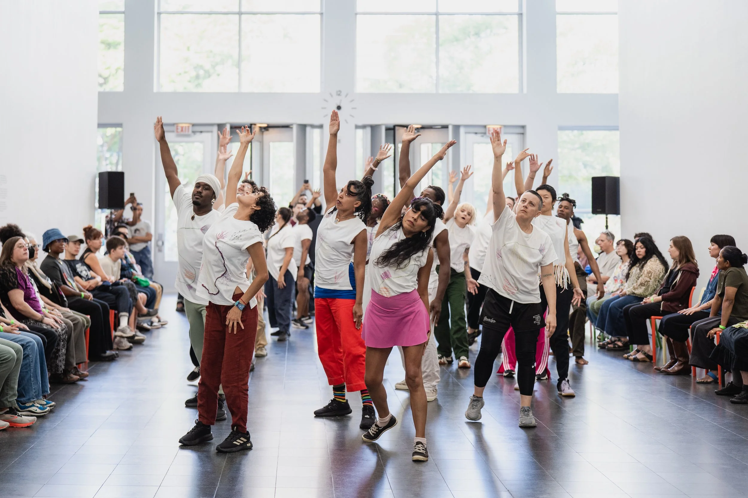 A group of performance facilitators cluster in a large hall wearing white t-shirts and plethora of colored bottoms.