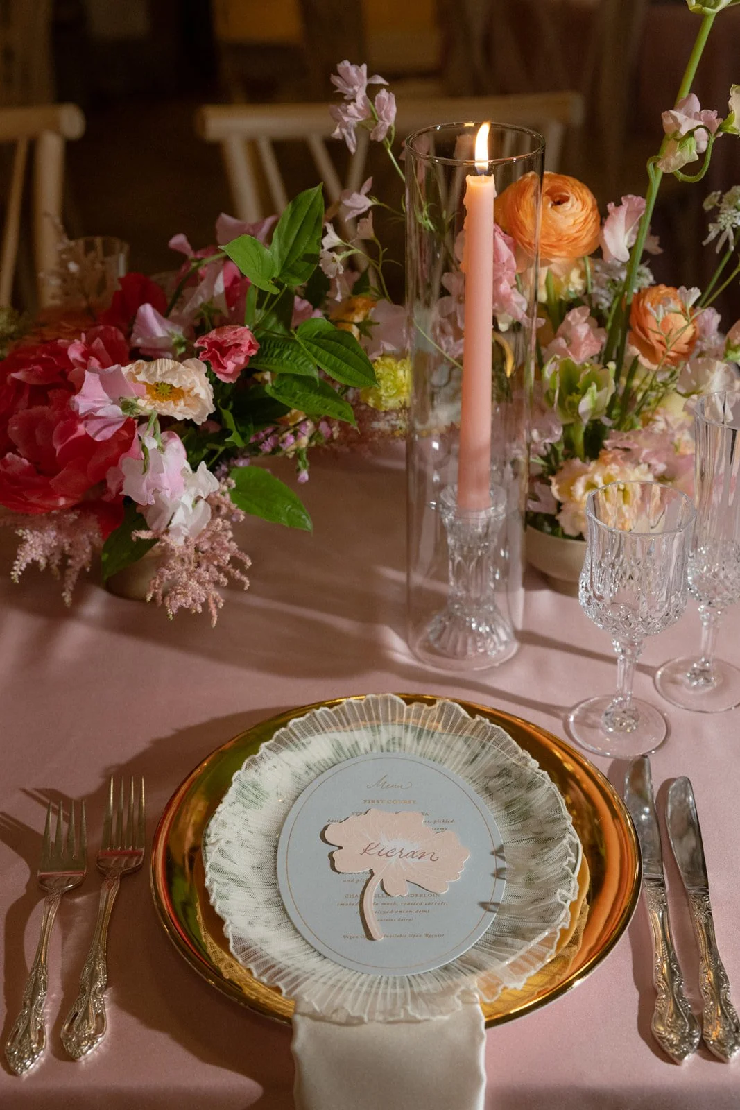 Elegant table setting with a floral centerpiece, a tall pink candle in a glass holder, crystal glassware, and a gold-rimmed plate with a menu card that has a pink leaf with the name 'Kieran' on it.