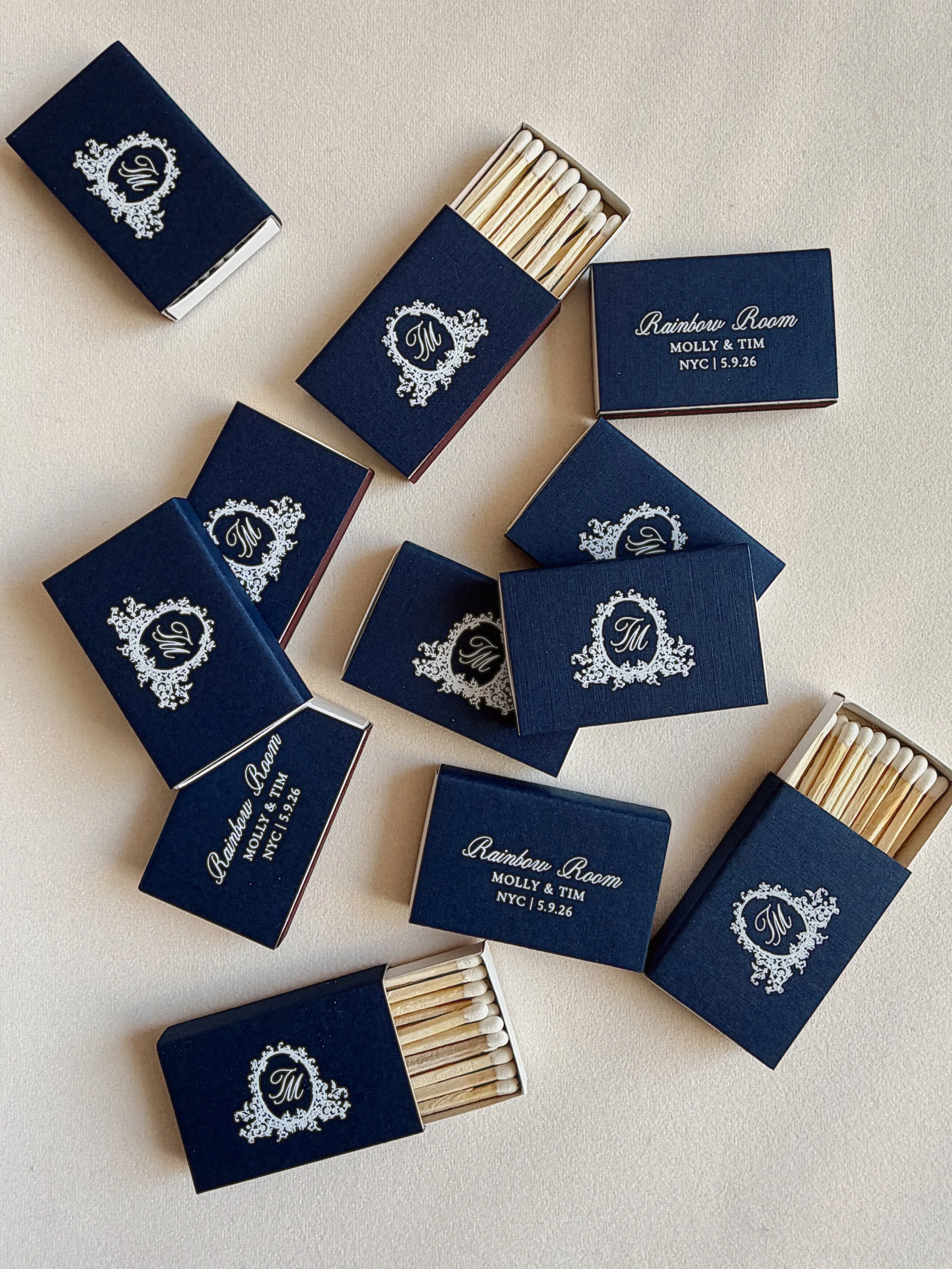 Several blue matchboxes with white ornate designs and text, several of which are open and containing wooden matches. One matchbox displays the text "Rainbow Room MOLLY & TIM NYC 5.9.26".