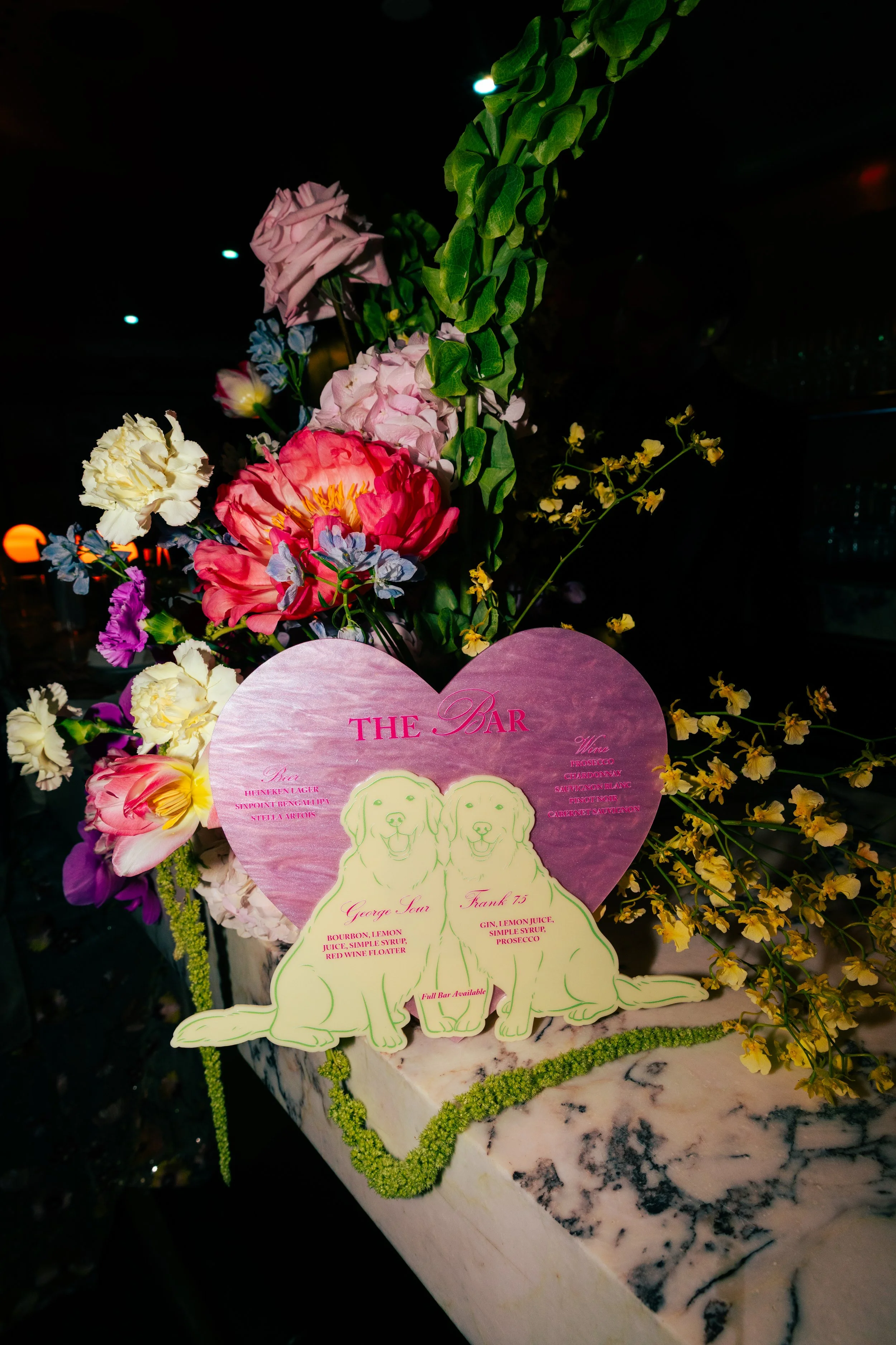 Colorful flower arrangement with pink roses, white, purple, blue, and yellow flowers, and green foliage. There is a pink heart-shaped sign in the front with a list of drinks and ingredients, including gin, lemon juice, simple syrup, and prosecco, alo