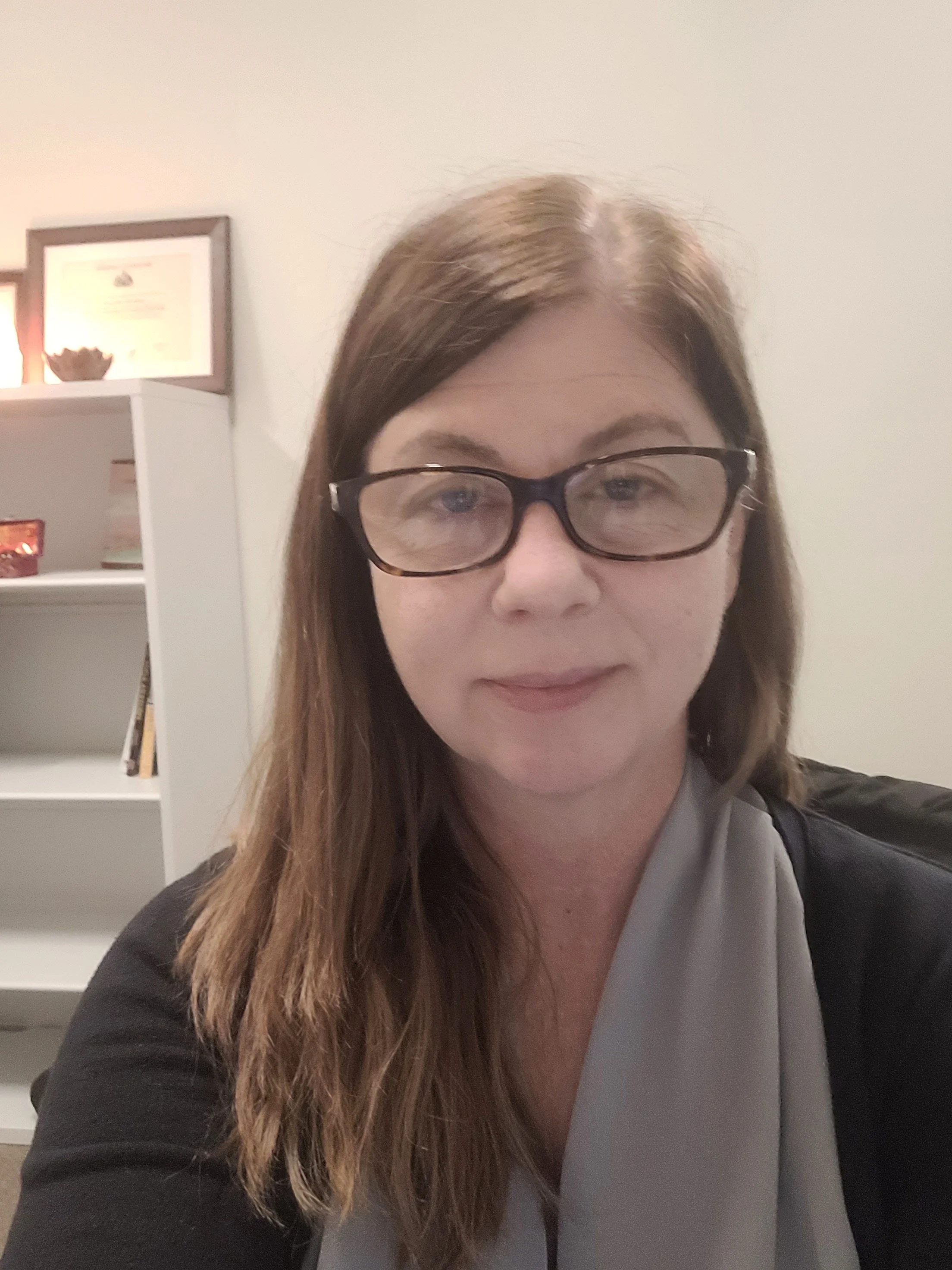 Close-up of a woman with long brown hair and black glasses, wearing a black jacket and gray shirt, in an indoor setting with white walls and a white bookshelf in the background.
