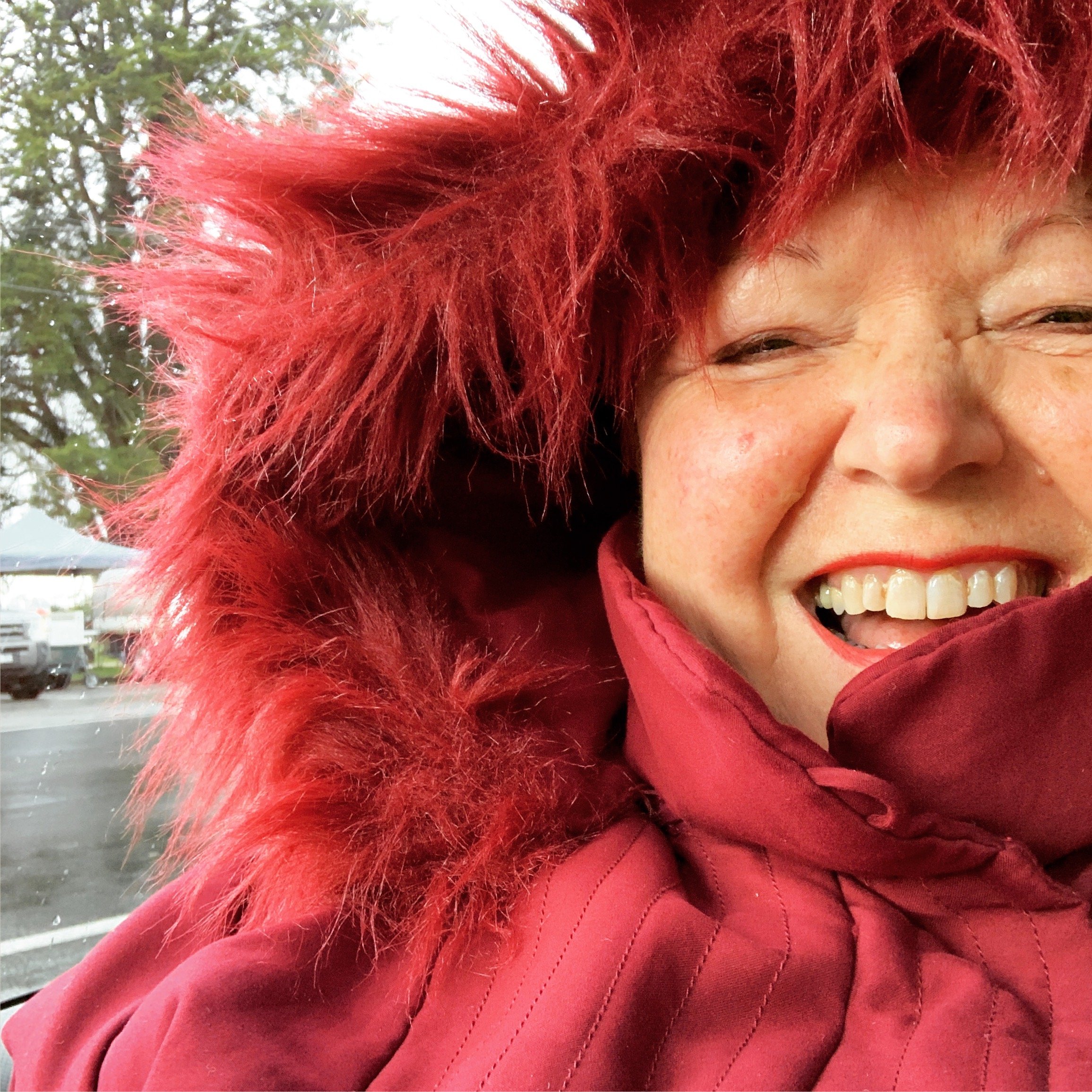 Close-up selfie of Janette, wearing a red jacket with a furry red hood, laughing with closed eyes. The background is a park full of trees, and it's raining.