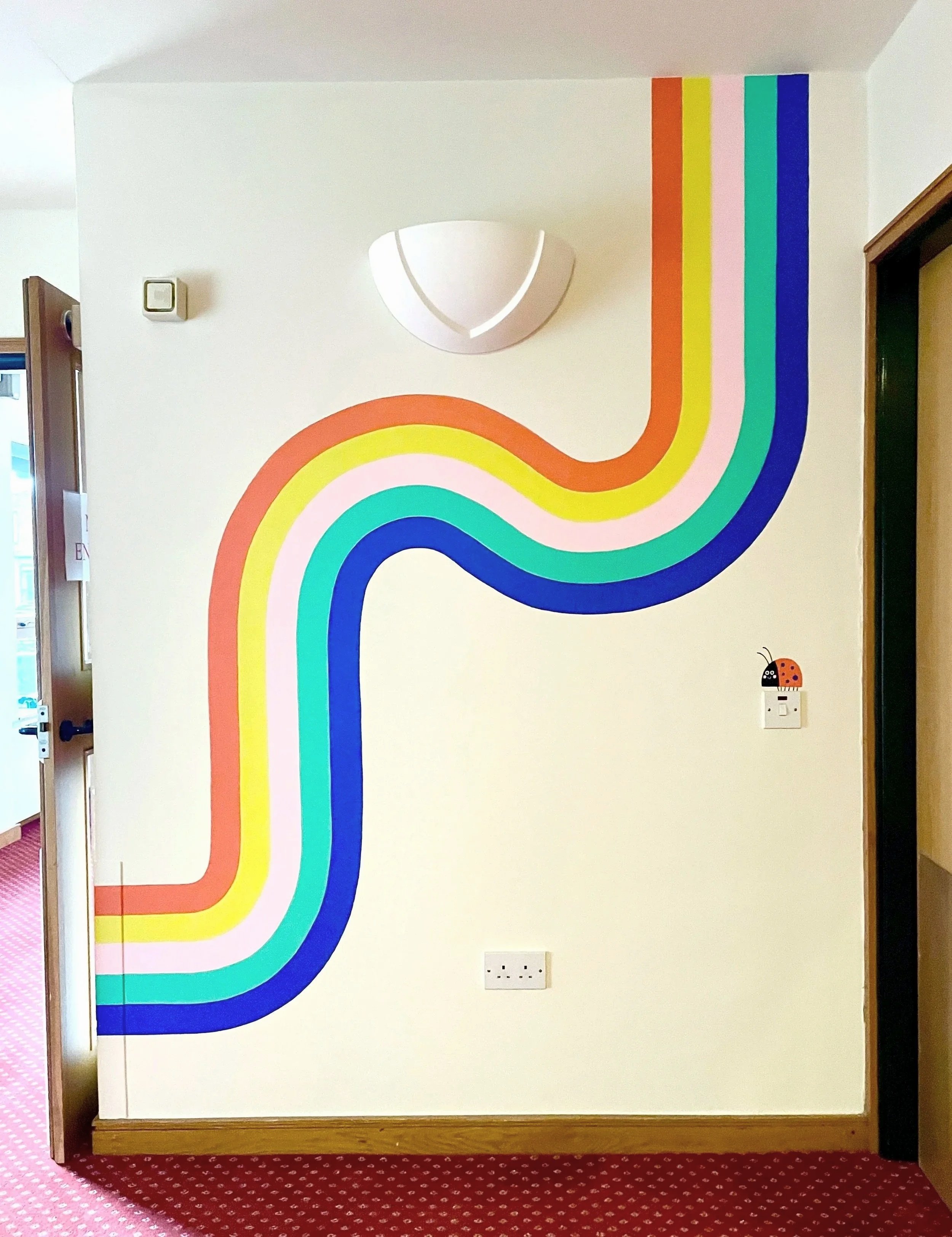 Section of corridor wall at Francis House hospice with hand painted rainbow wave mural plus a ladybird on the light switch, by artist Melanie Williamson