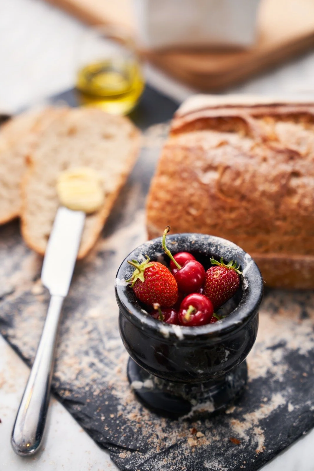 A portrait view of a baking scene, with fresh strawberries and cherries in a black marble pot in the foreground.