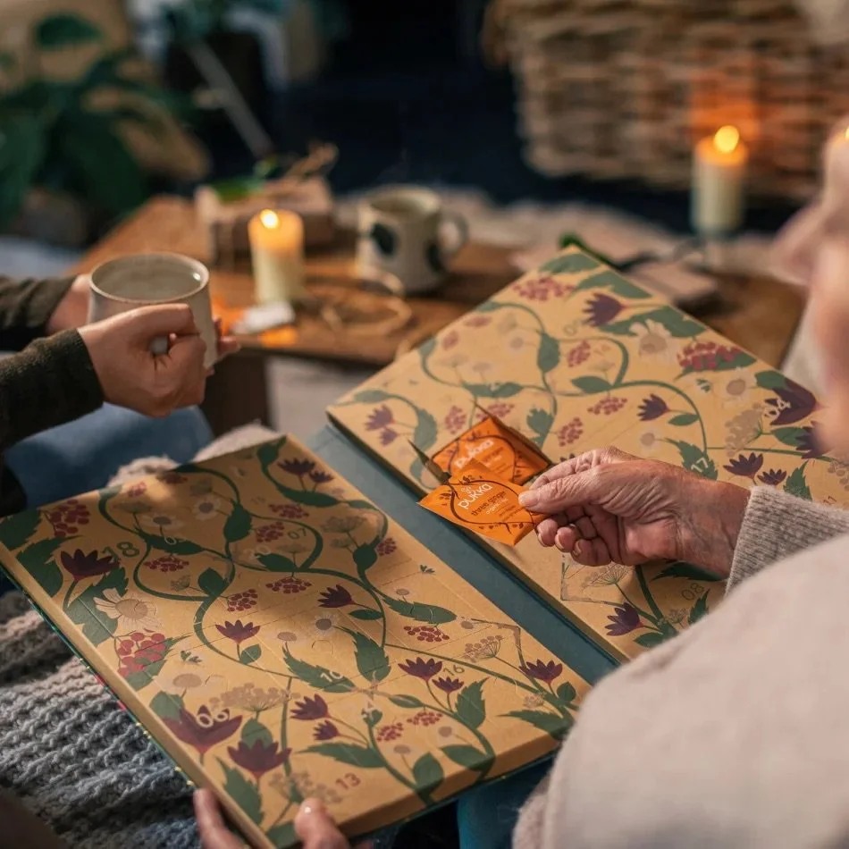 Close-up of a person opening a Christmas advent calendar with numbered compartments and a Christmas-themed design, surrounded by candles, a mug and a teapot on a cozy wooden table during a festive gathering.