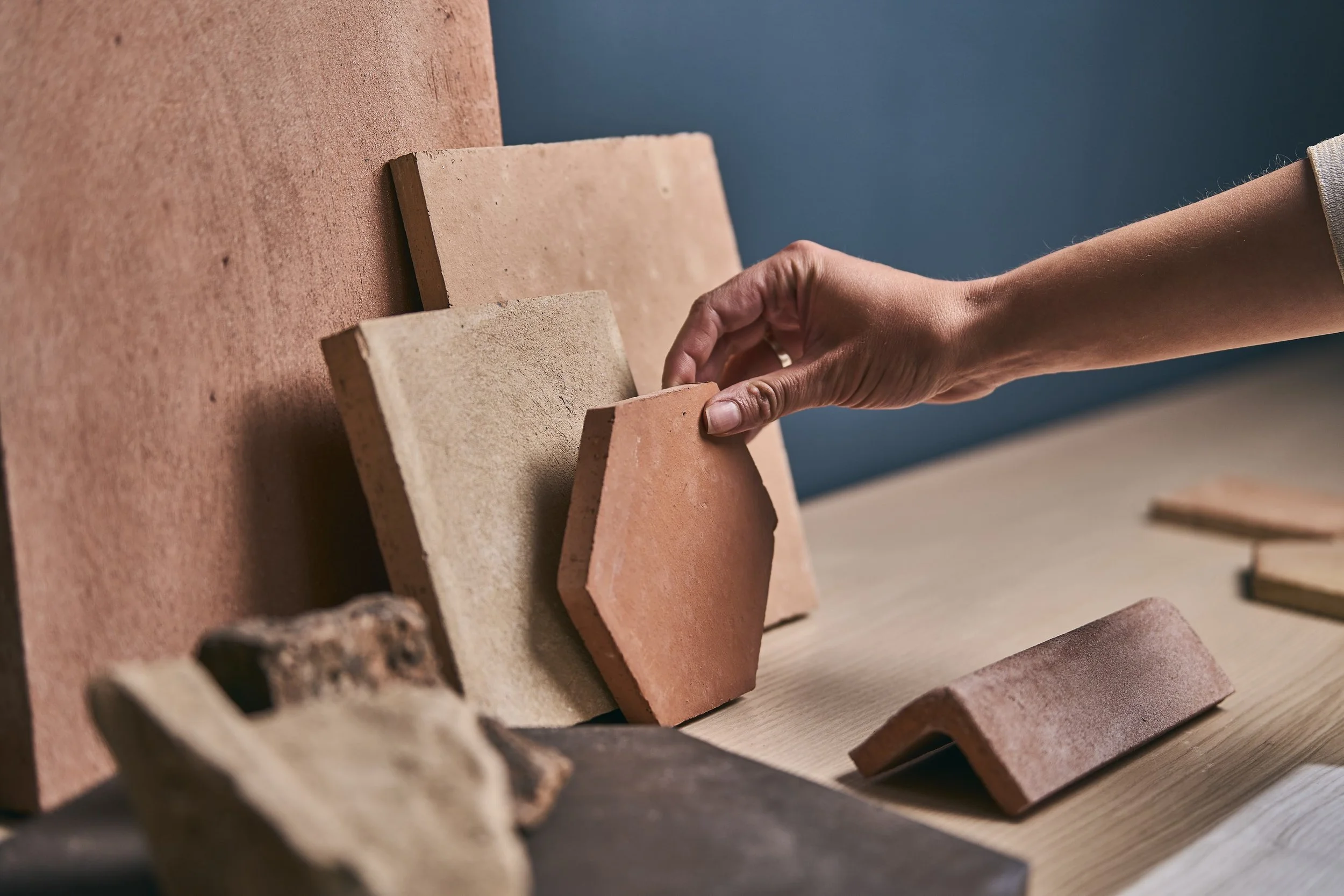 A hand reaching out to pick up a terracotta tile from a display of various bricks on a wooden surface.