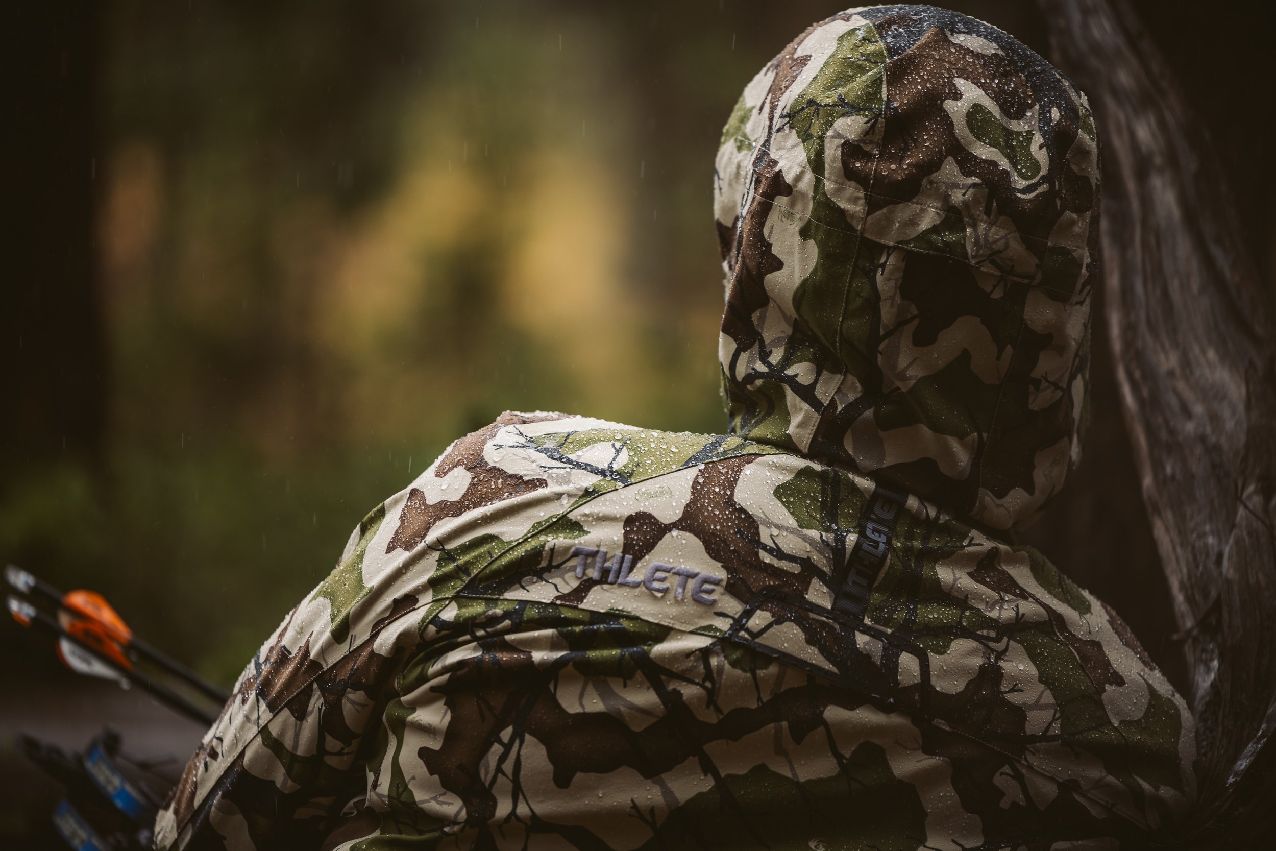 Person wearing a camouflage-patterned rain jacket with a hood, holding arrows, in a forest setting.