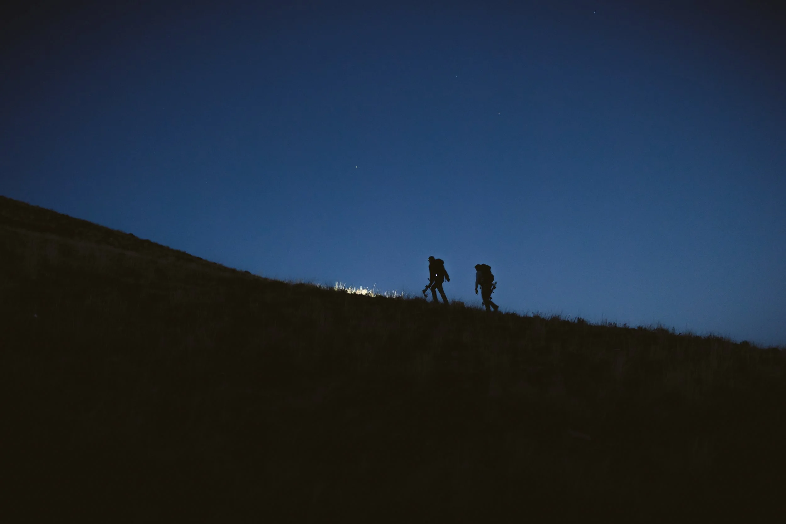 Silhouettes of two hikers with backpacks walking uphill at night, with a clear blue sky in the background.