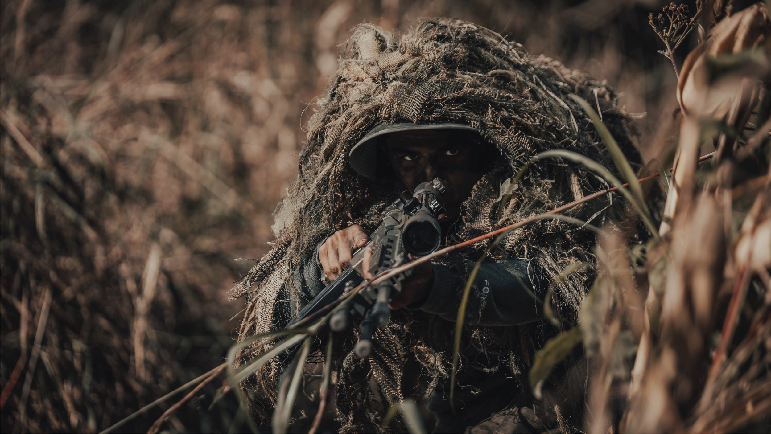Sniper in camouflage aiming through rifle scope, hidden in tall grass.