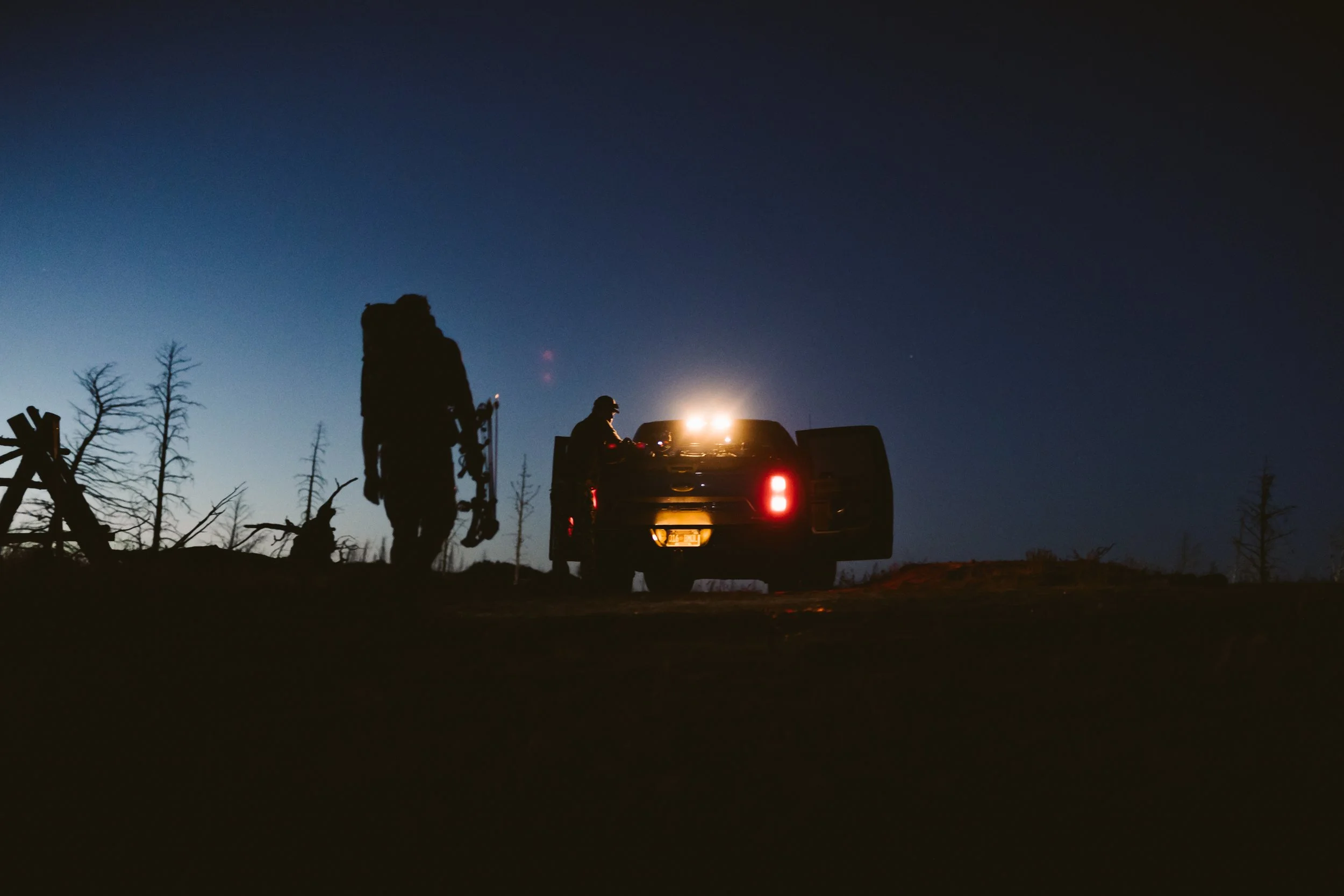 Silhouette of a person near a parked truck with headlights on at dusk, surrounded by leafless trees.