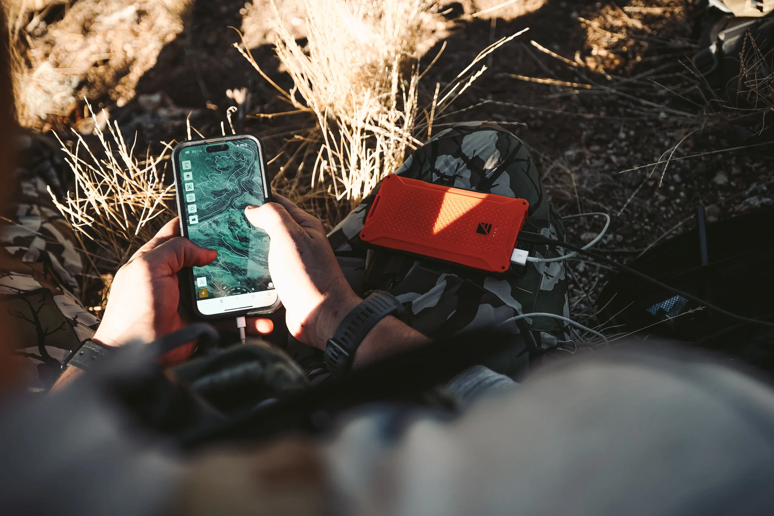 Person using smartphone with topographic map outdoors, portable charger nearby.