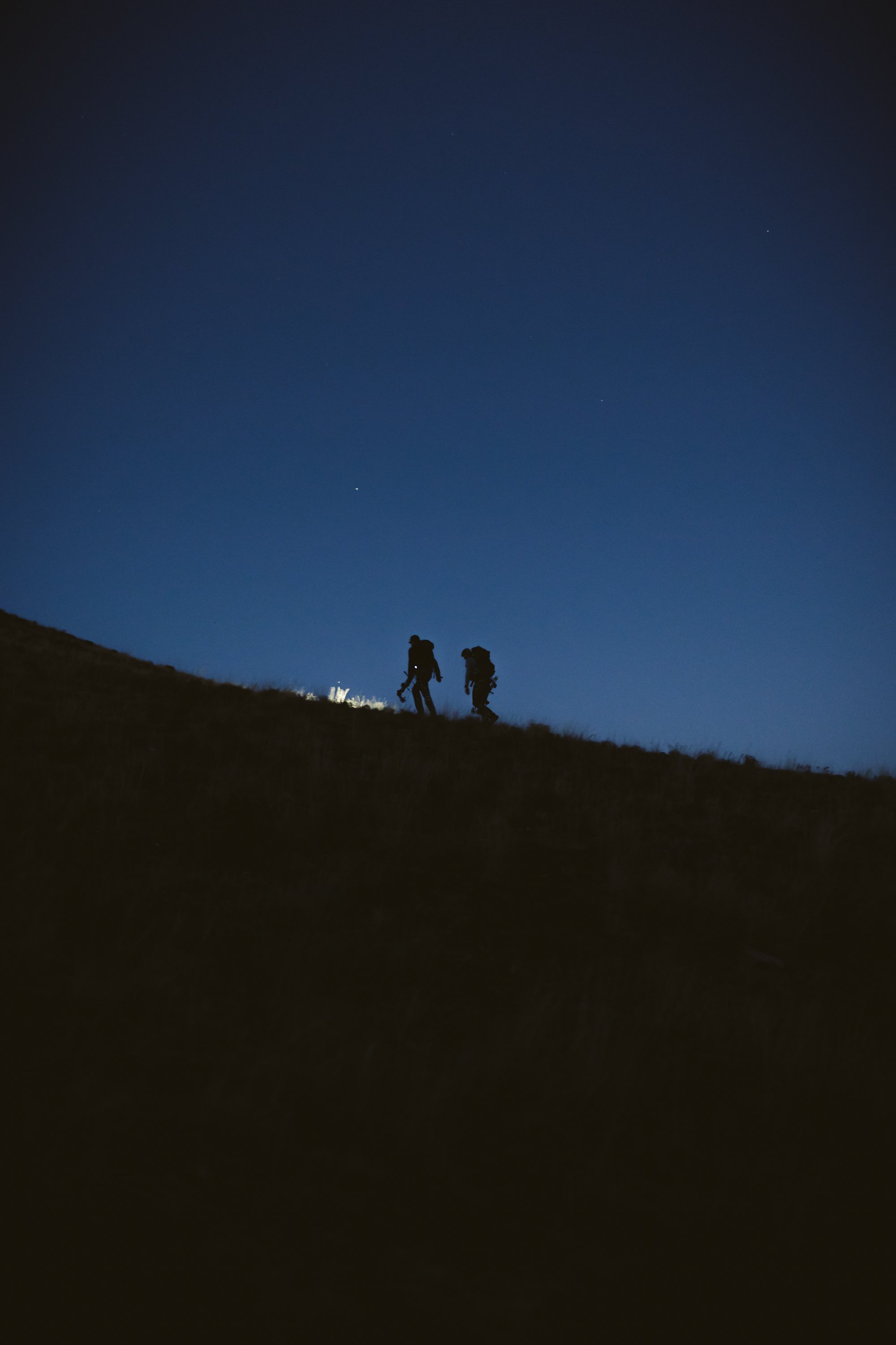 Silhouettes of two hikers with backpacks walking uphill under a clear night sky.