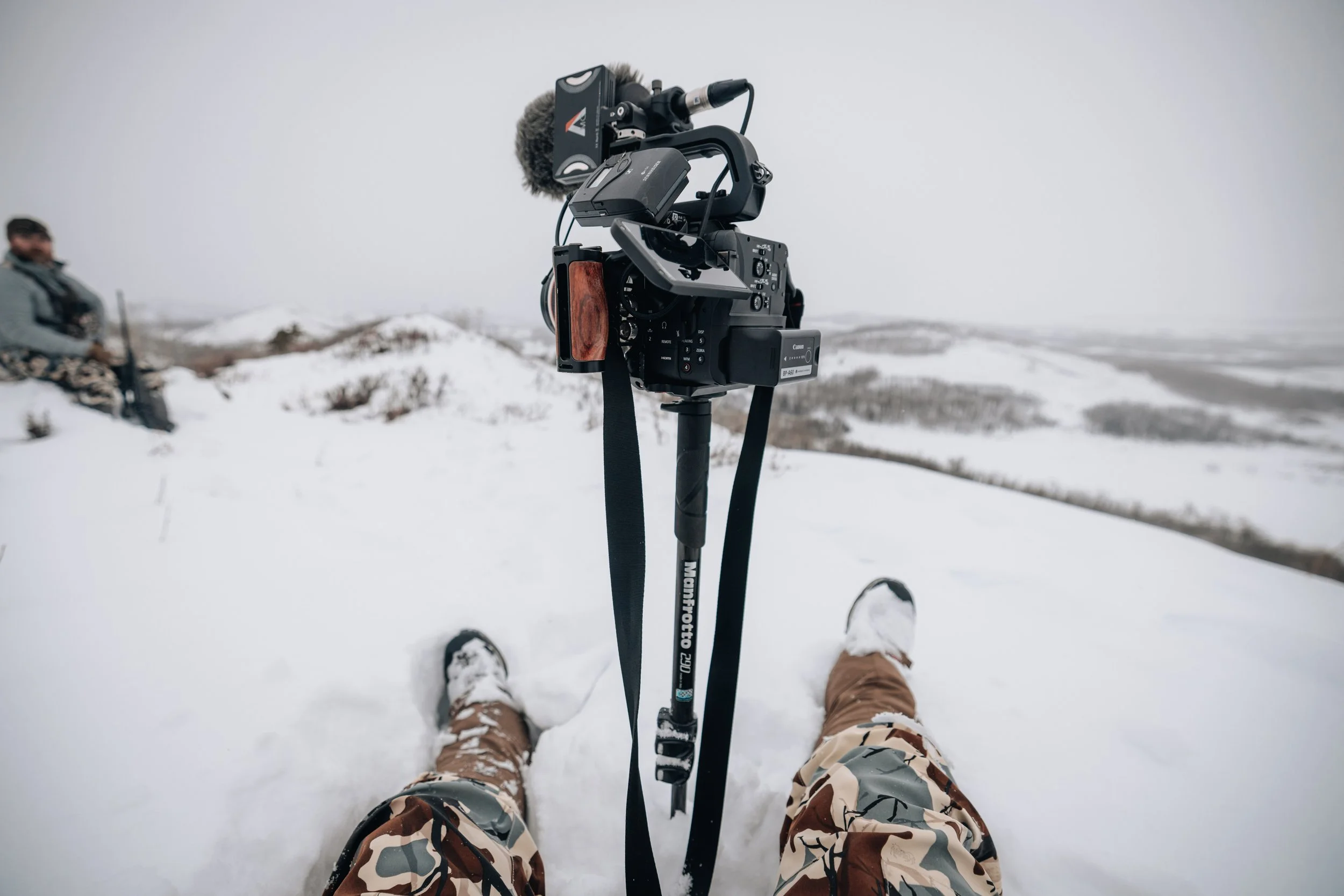 Person in snow with camo pants and a camera on a tripod in winter landscape.