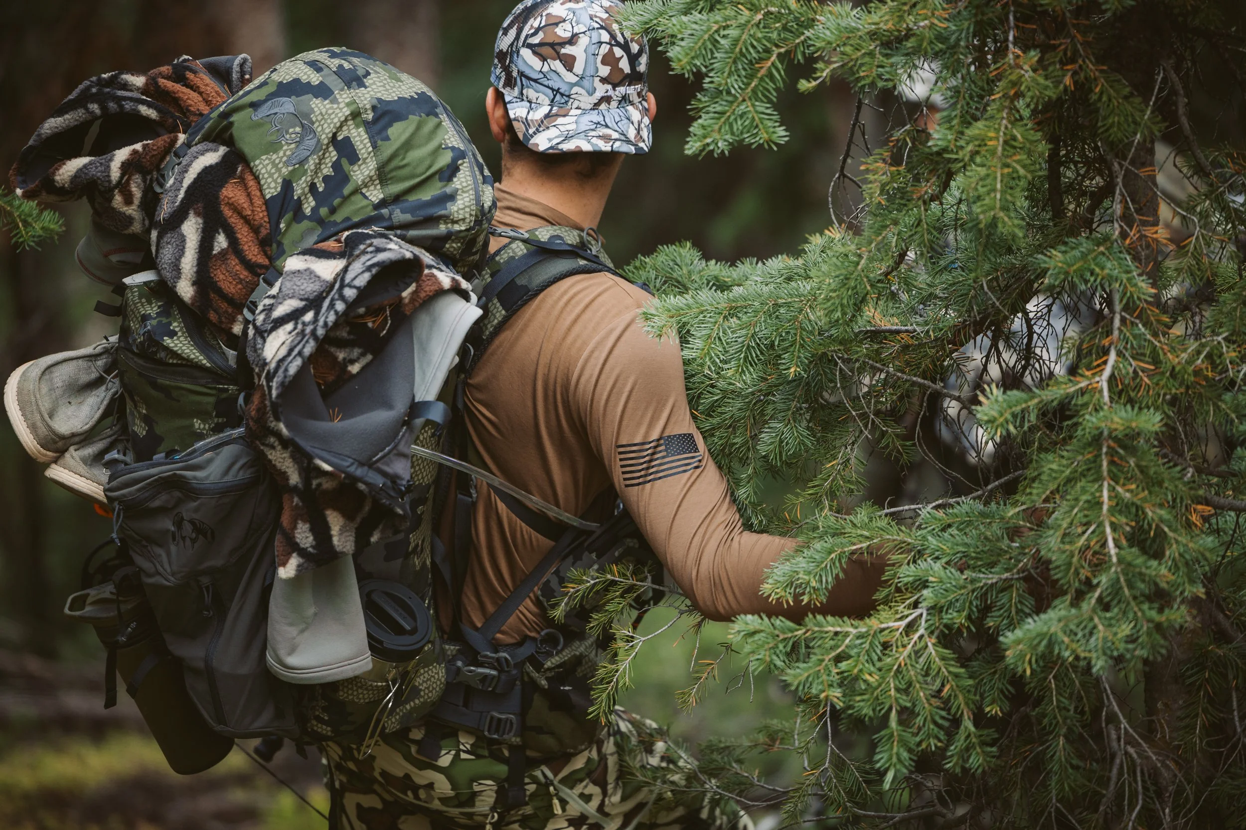 Person in camouflage gear with a large backpack walking through a forest, partially obscured by evergreen trees.