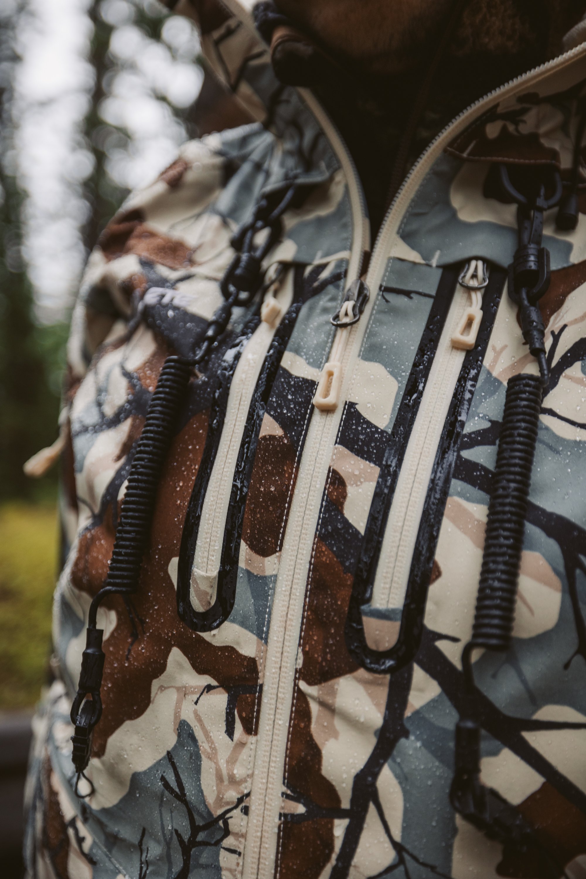 Close-up of a camouflage rain jacket in a forest setting, with visible water droplets on the surface.