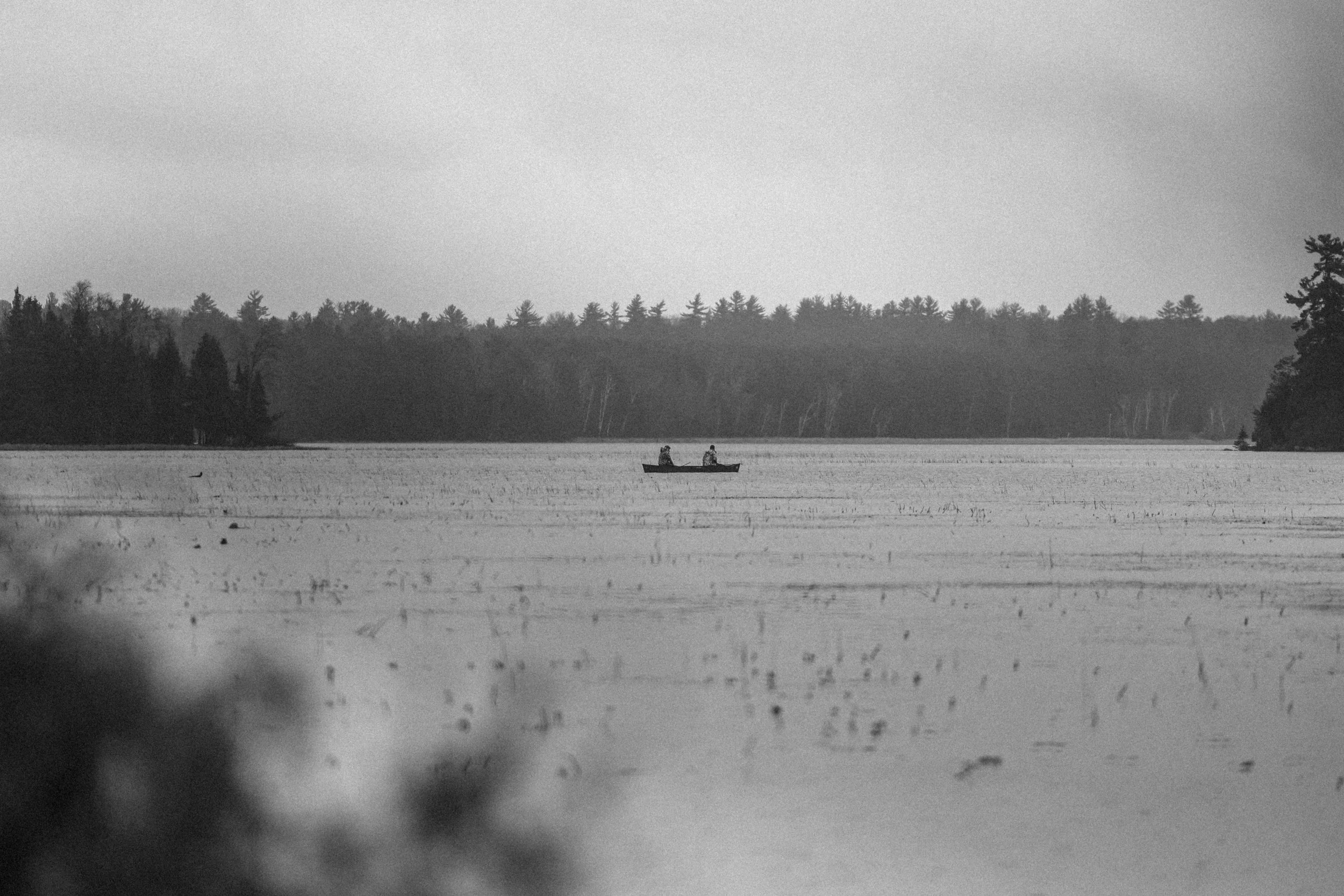 Black and white photo of a canoe with two people on a large lake, surrounded by forest trees in the background.