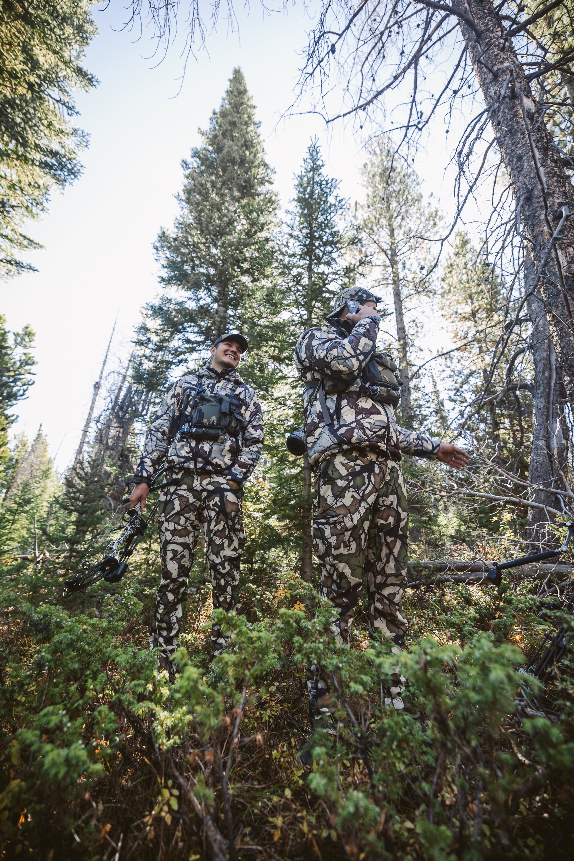 Two people in camouflage clothing standing in a dense forest, one holding a bow.