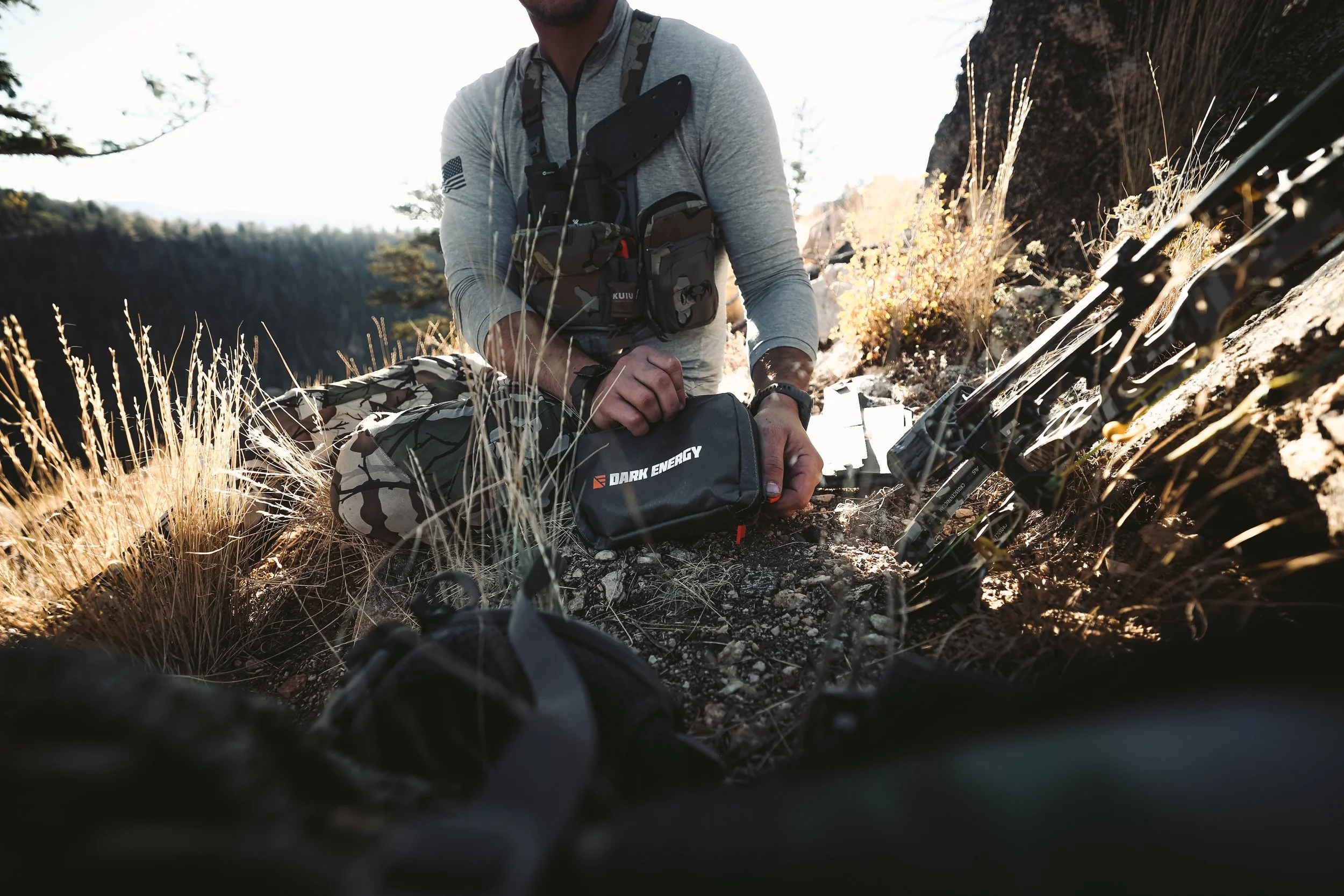 Person wearing camo and gray shirt sitting on rocky ground holding a Dark Energy pouch, with outdoor gear around.