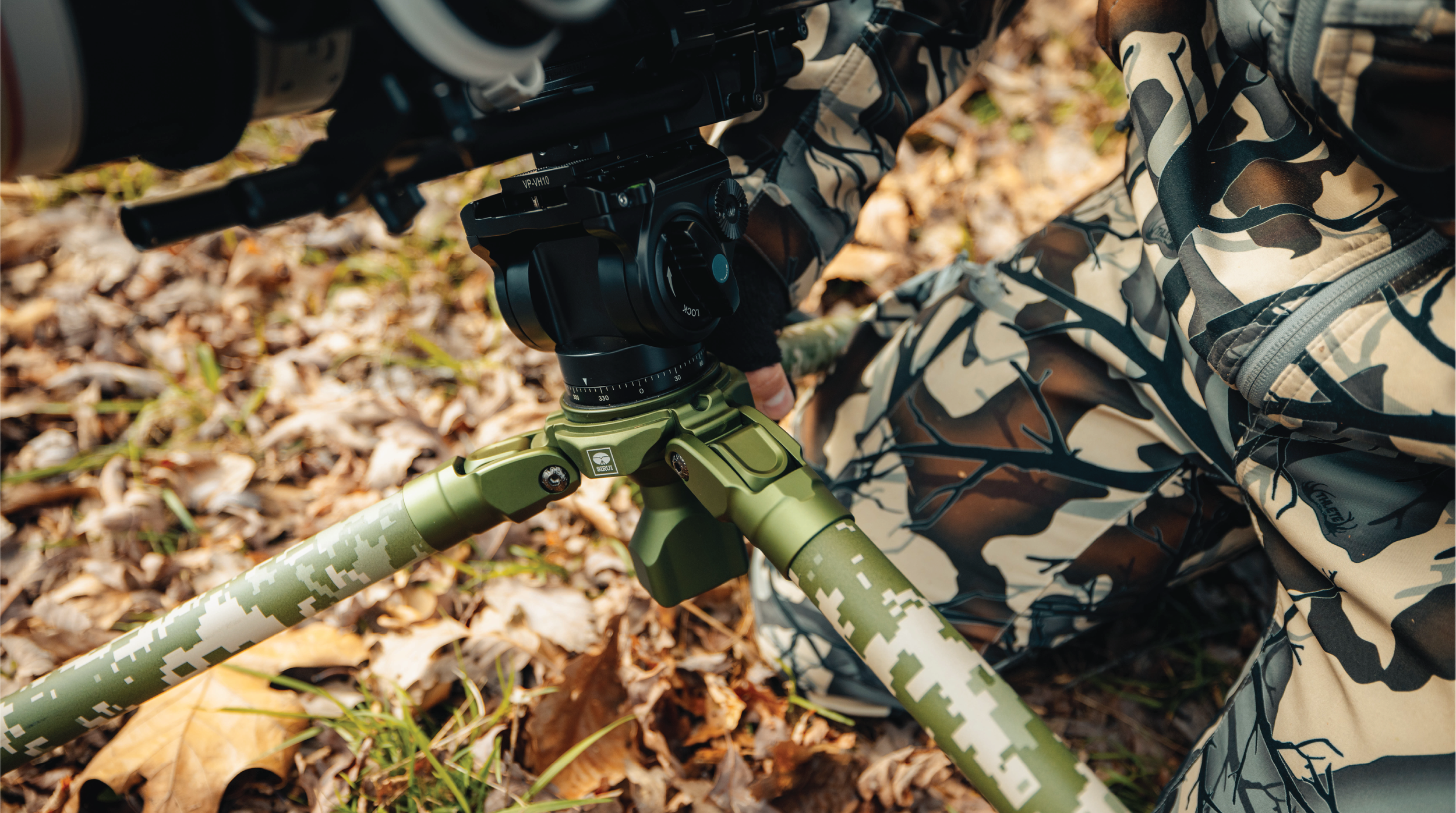 Close-up of a camera tripod with green digital camouflage legs, and a person wearing camouflage clothing on a forest floor with dried leaves.