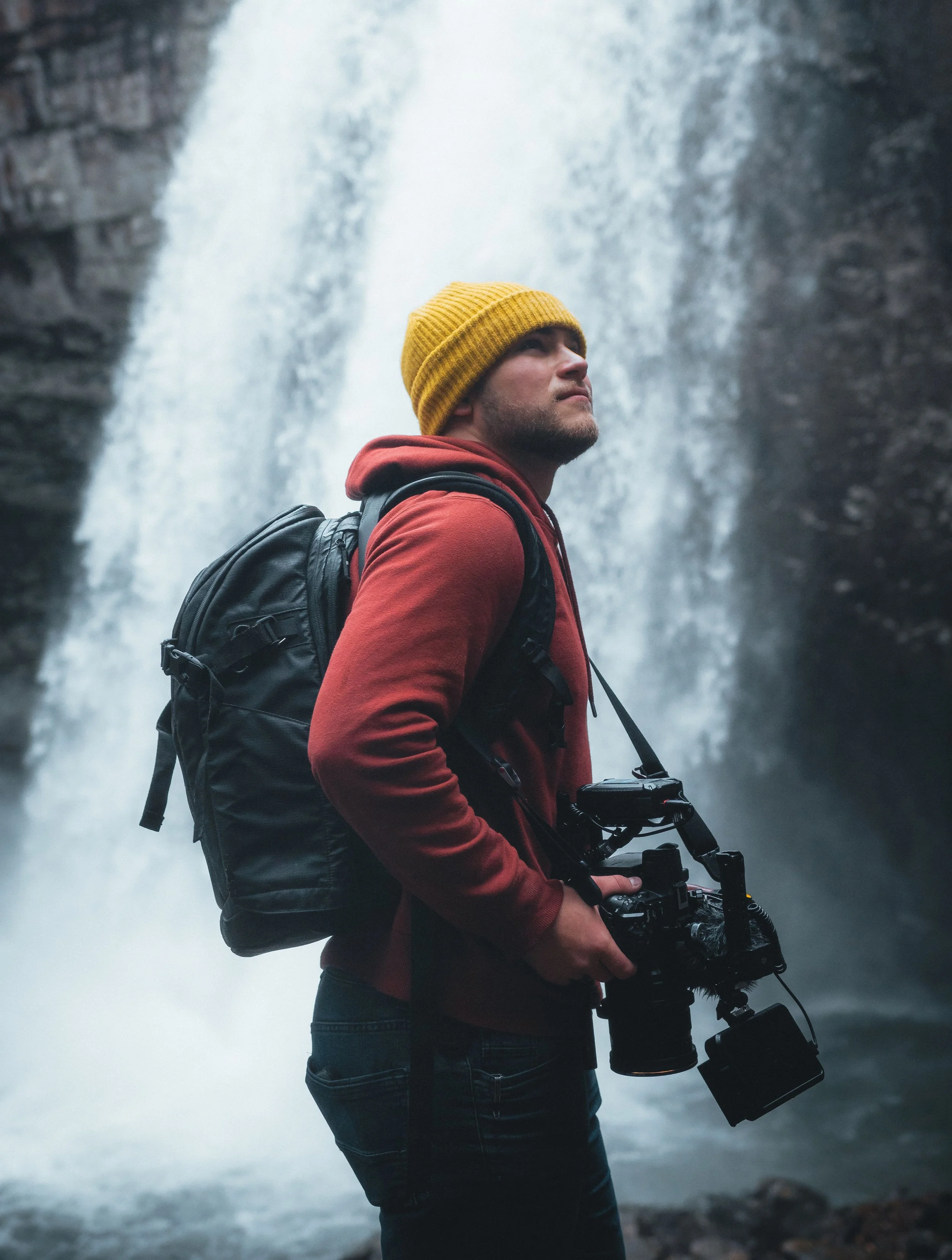 Man in a red hoodie and yellow beanie with a backpack and camera standing in front of a waterfall.