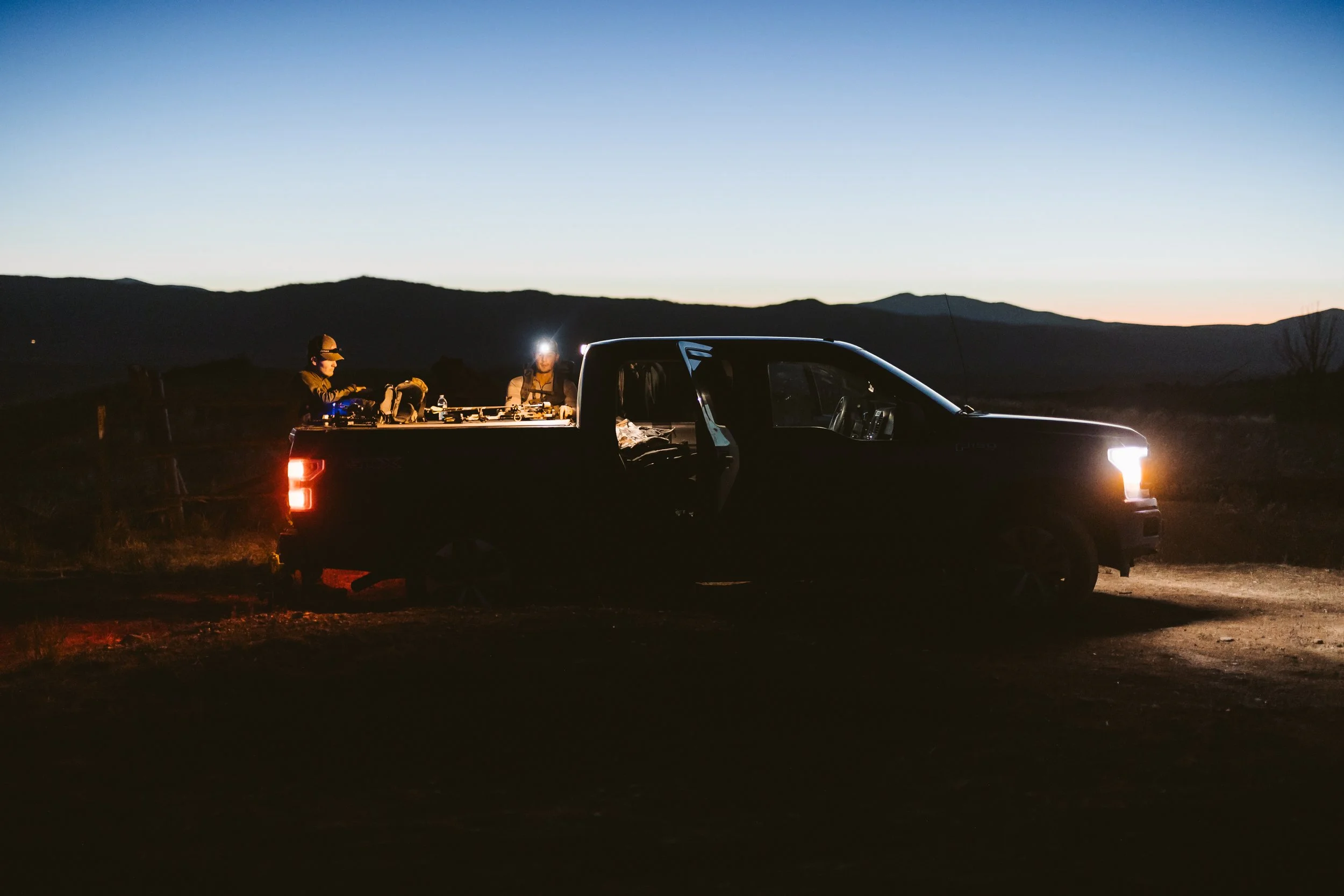 Pickup truck with open doors at sunset, people working in truck bed, mountainous background.