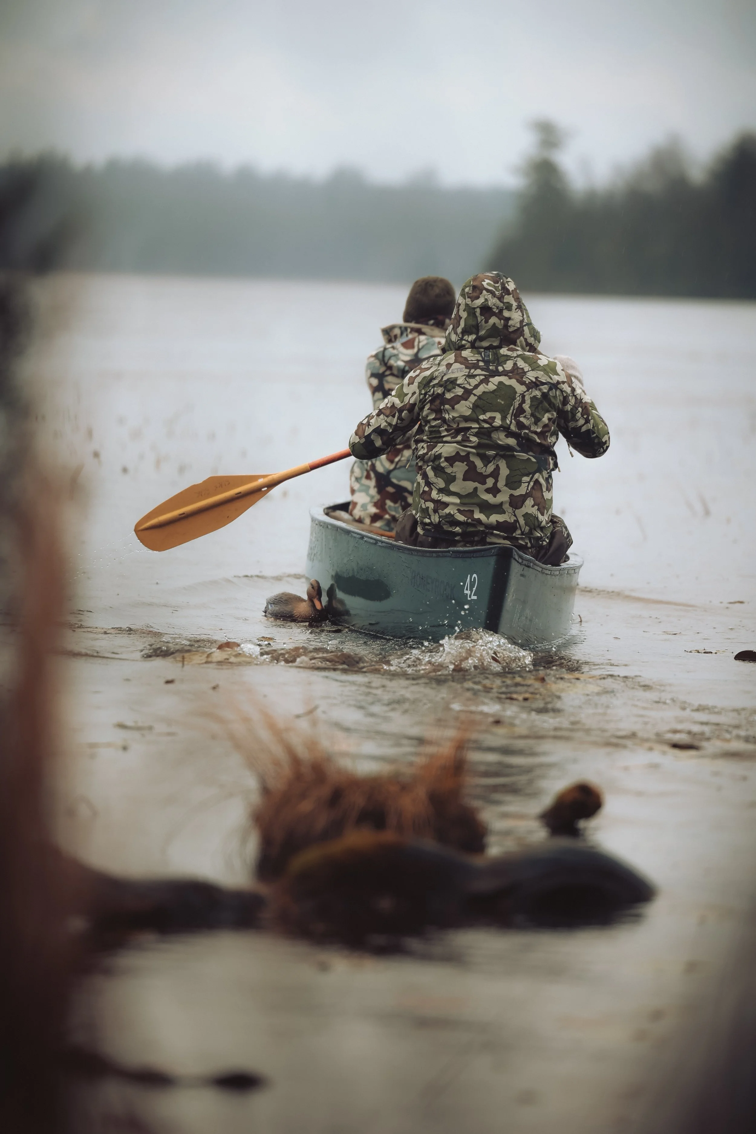 Two people in camouflage jackets paddling a canoe on a lake, surrounded by water and distant trees.