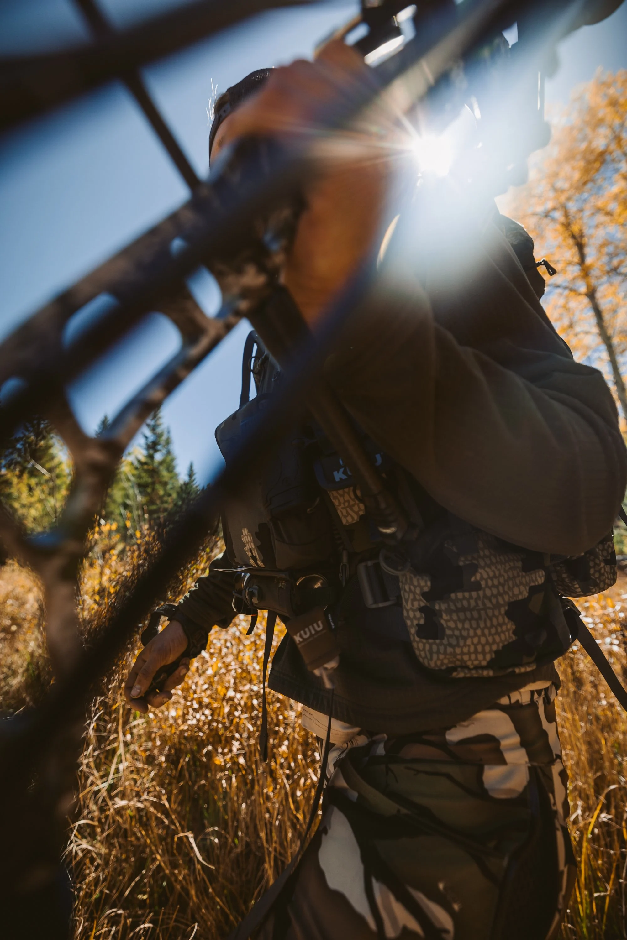 Person in camouflage gear holding a compound bow outdoors with sunlight behind, surrounded by dry grass and trees.