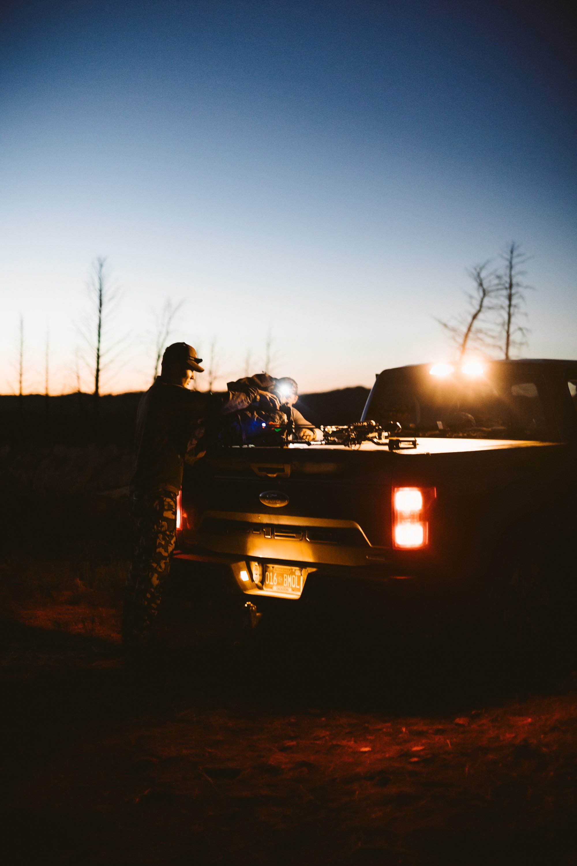 Two people standing at the open trunk of a pickup truck at dusk, with the truck's lights on, surrounded by a dimly lit outdoor setting.