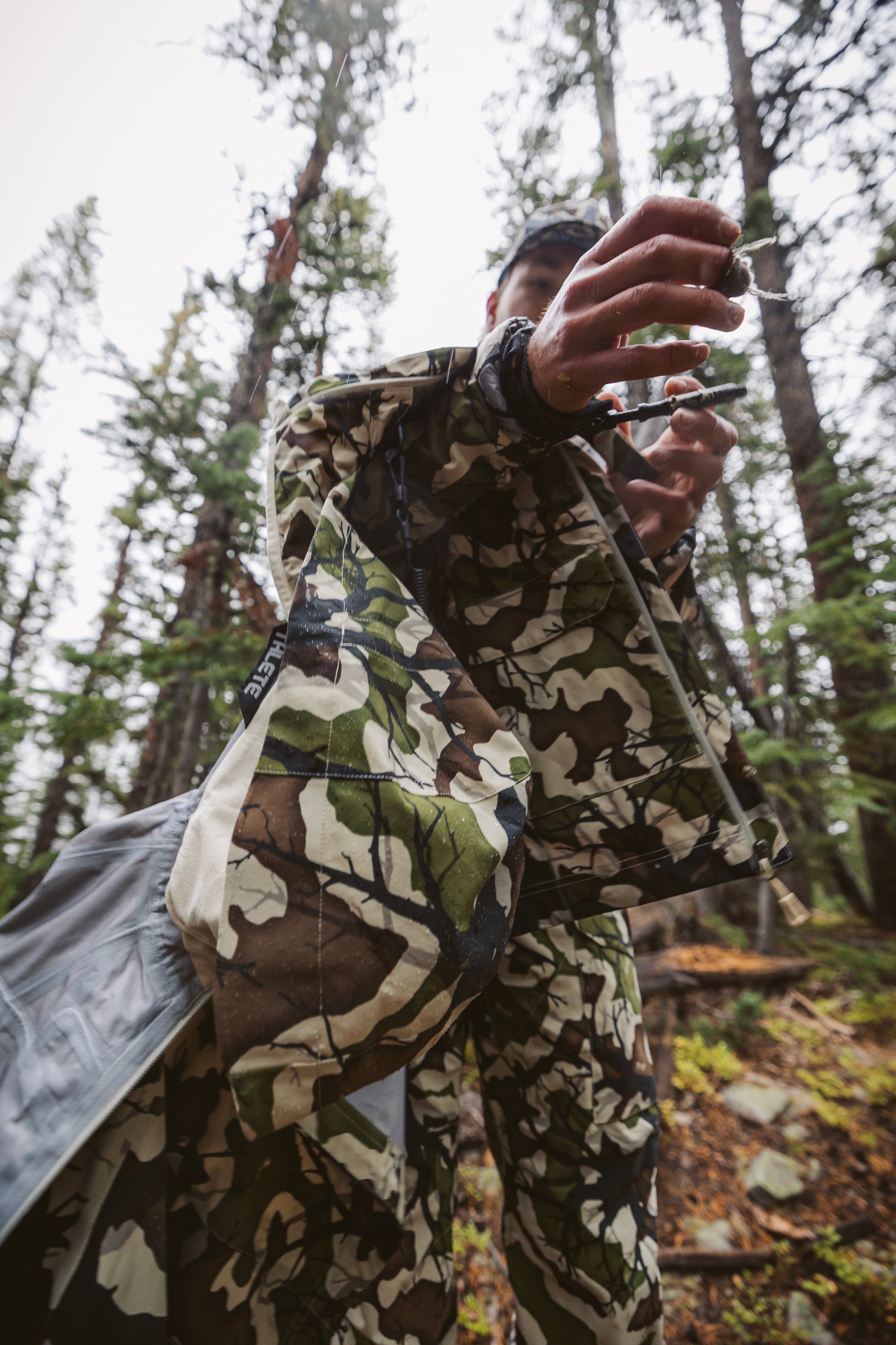Person wearing camouflage clothing outdoors in a forest, reaching towards the camera with a hand extended, surrounded by tall trees.