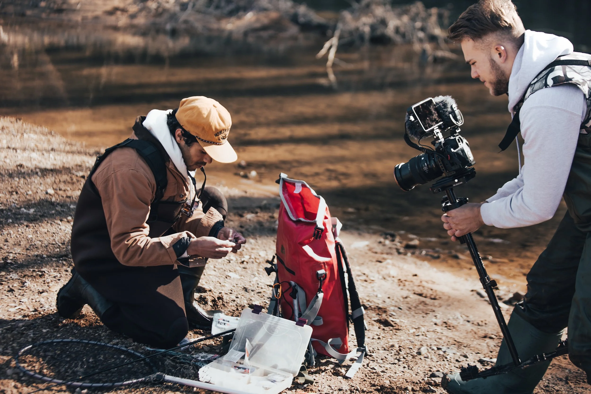 Person kneeling by a riverbank preparing fishing tackle while another person films with a camera on a tripod.