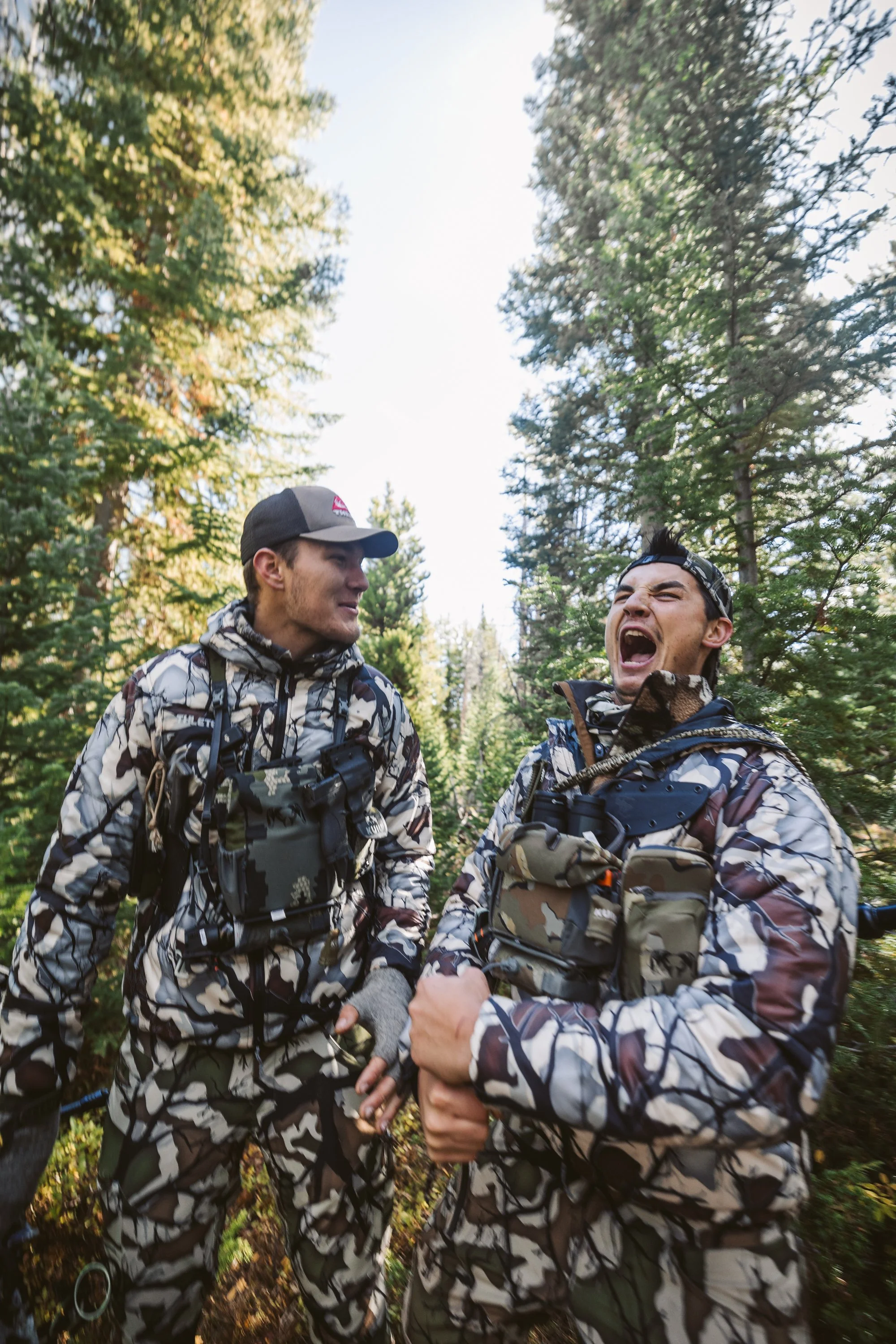 Two men in camouflage clothing laughing in a forest setting.