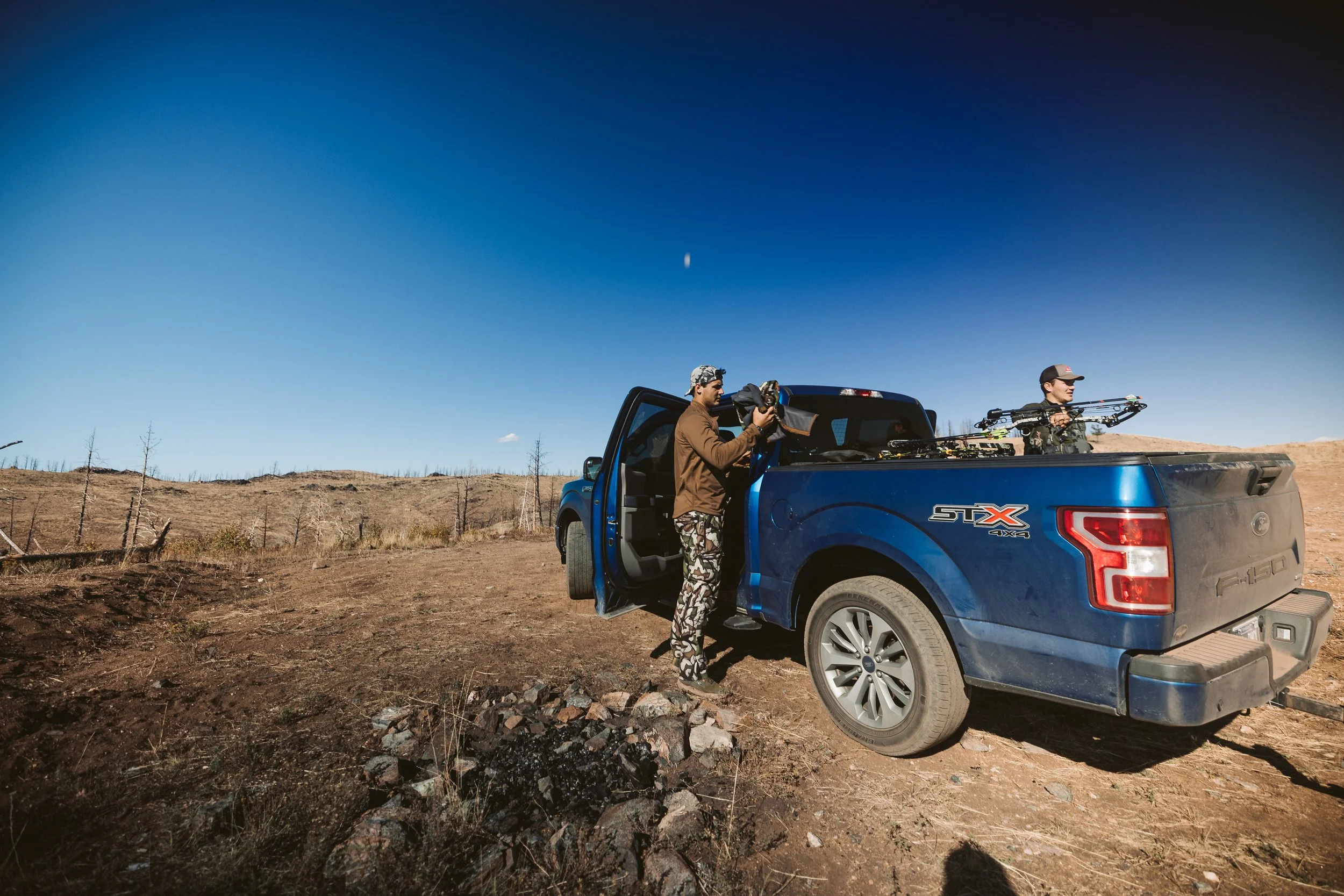 Two people standing near a blue Ford F-150 truck in a dry, open landscape. One person is at the open driver-side door, while the other stands in the truck bed holding a compound bow. The sky is clear and blue.